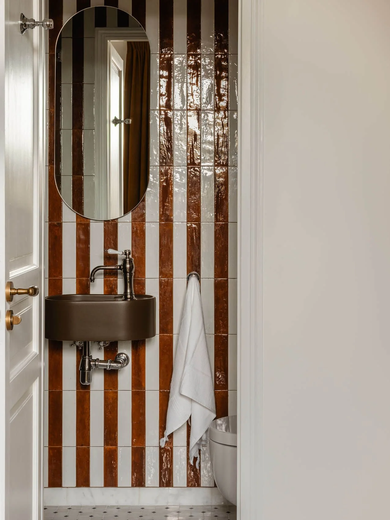 Paris apartment powder room renovation with striped ceramic wall tiles, wall-mounted sink, arched mirror, vintage-style brass hardware, and compact bathroom layout.