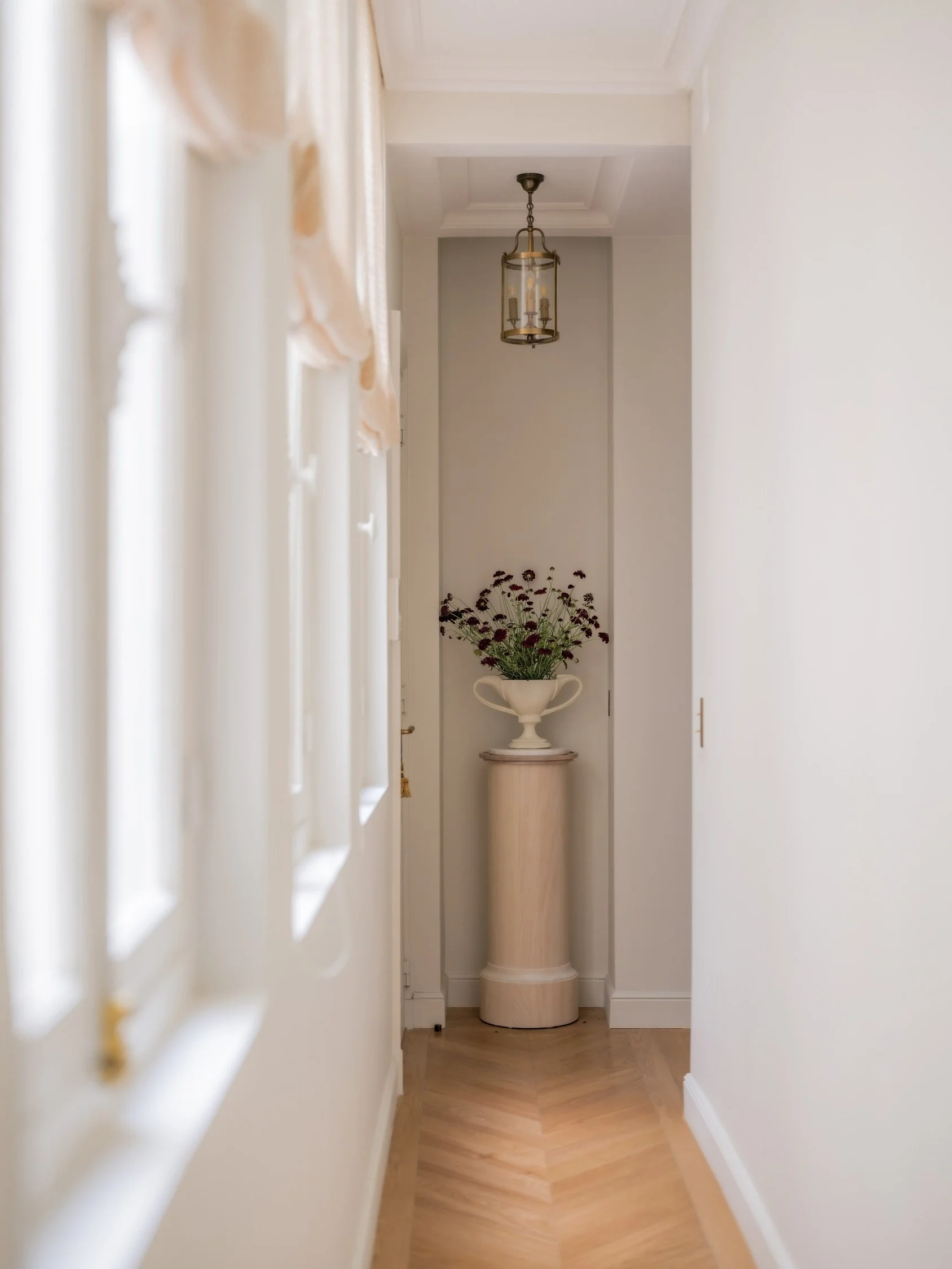 Parisian apartment hallway with herringbone parquet floor, stone pedestal with classical urn and wildflowers, and antique brass lantern pendant