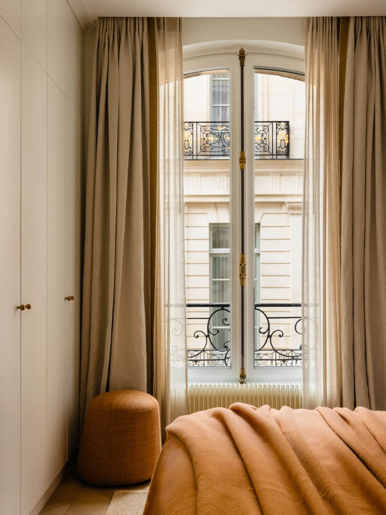 Paris bedroom renovation detail with arched French window, sheer and blackout curtains, built-in wardrobe cabinetry, textured ottoman, and soft layered bedding.
