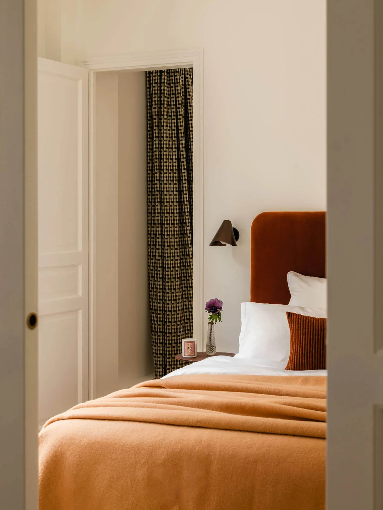 Paris apartment bedroom renovation with rust velvet headboard, layered white and terracotta bedding, wall-mounted reading light, and patterned accent curtains.