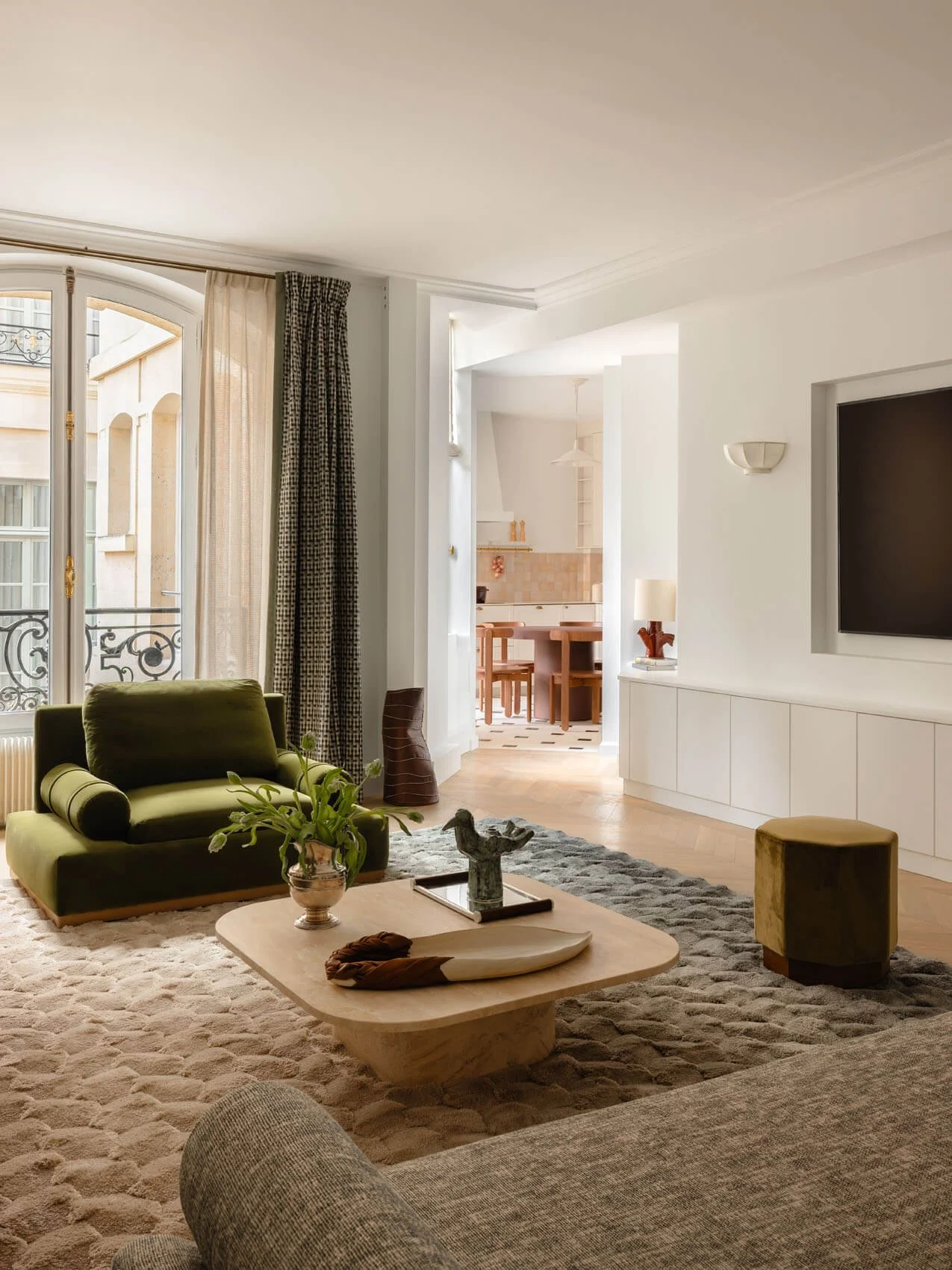 Open-plan Paris apartment living room renovation with green velvet seating, sculptural coffee table, built-in media wall, and view into modern kitchen and dining area.