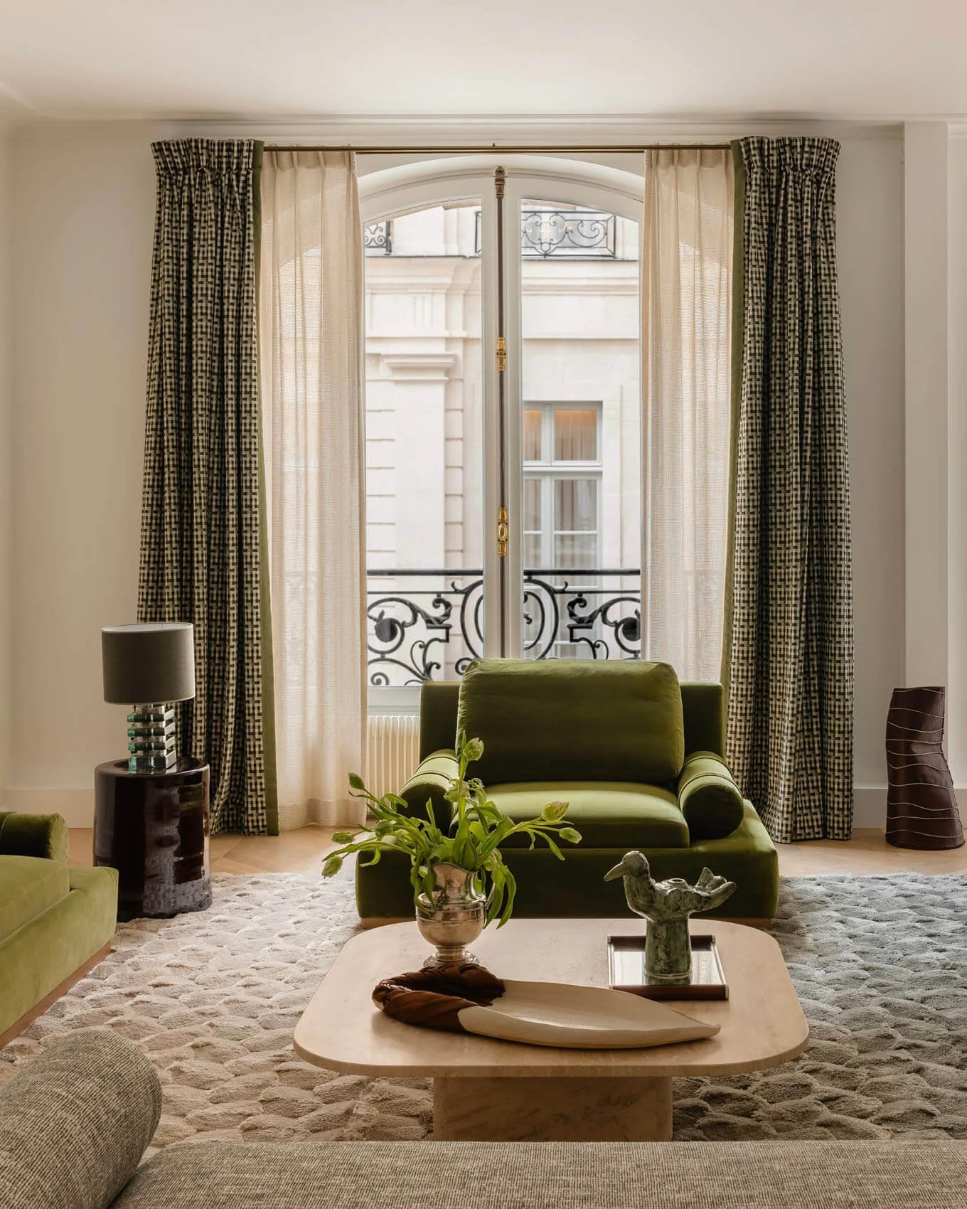 Paris living room renovation with green velvet armchair, custom curtains, sculptural coffee table, textured rug, and classic French window overlooking wrought-iron balcony.