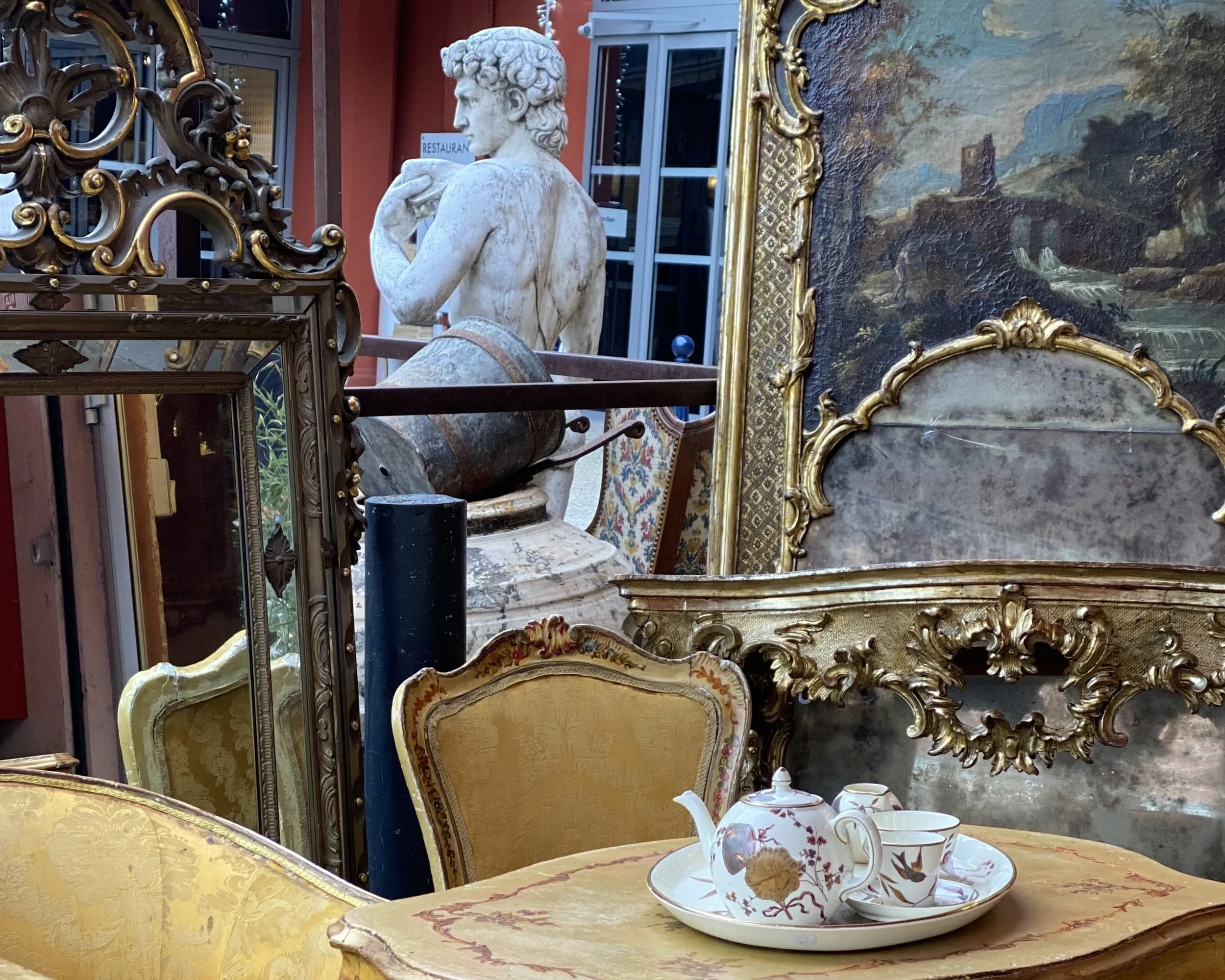 Antique gilded mirrors, a painted landscape, yellow damask chairs and a porcelain tea set on a painted table at Les Puces, Paris