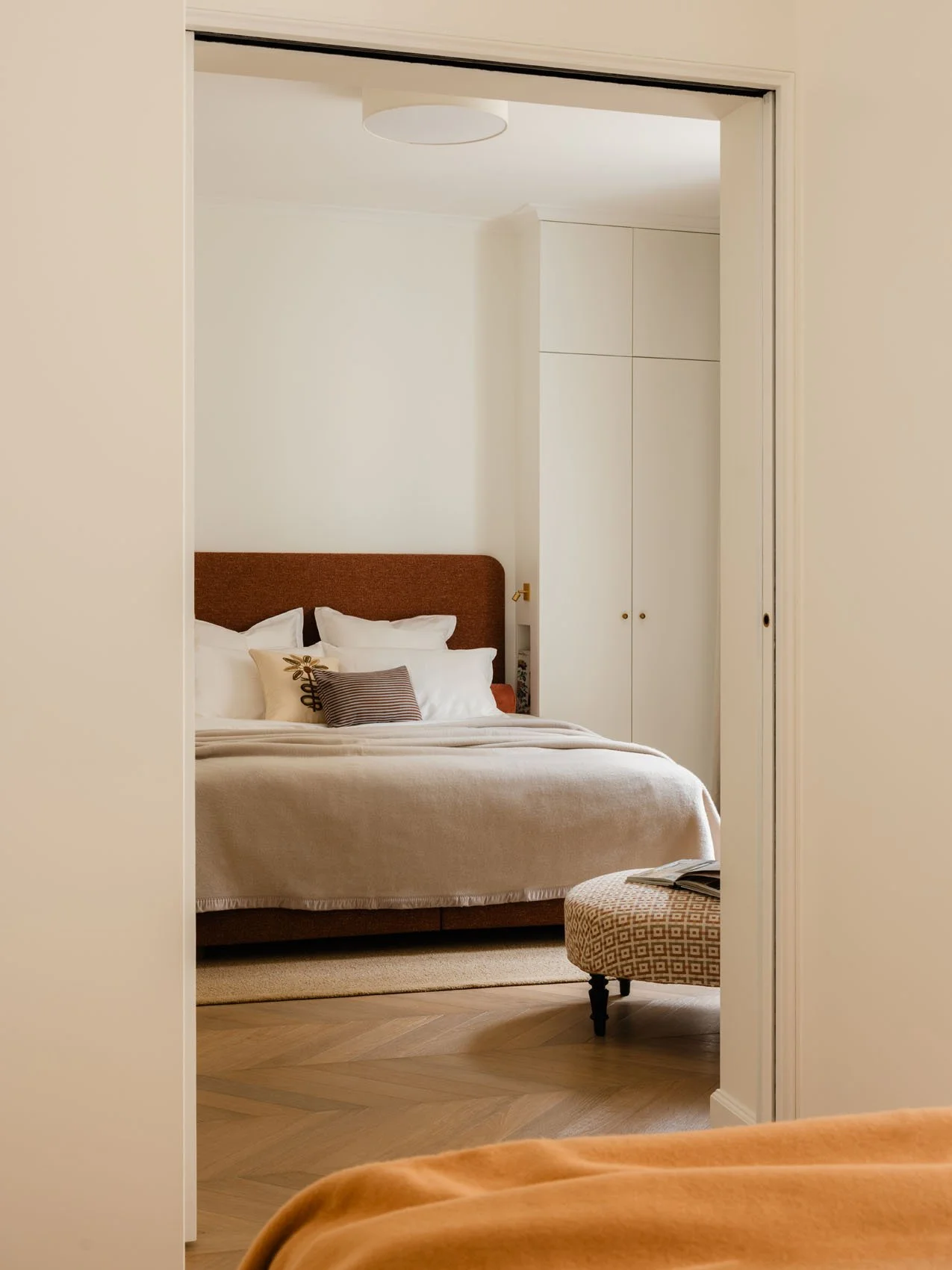 Paris apartment bedroom renovation viewed through sliding doorway, featuring upholstered headboard, layered neutral bedding, custom wardrobes, and herringbone wood flooring.