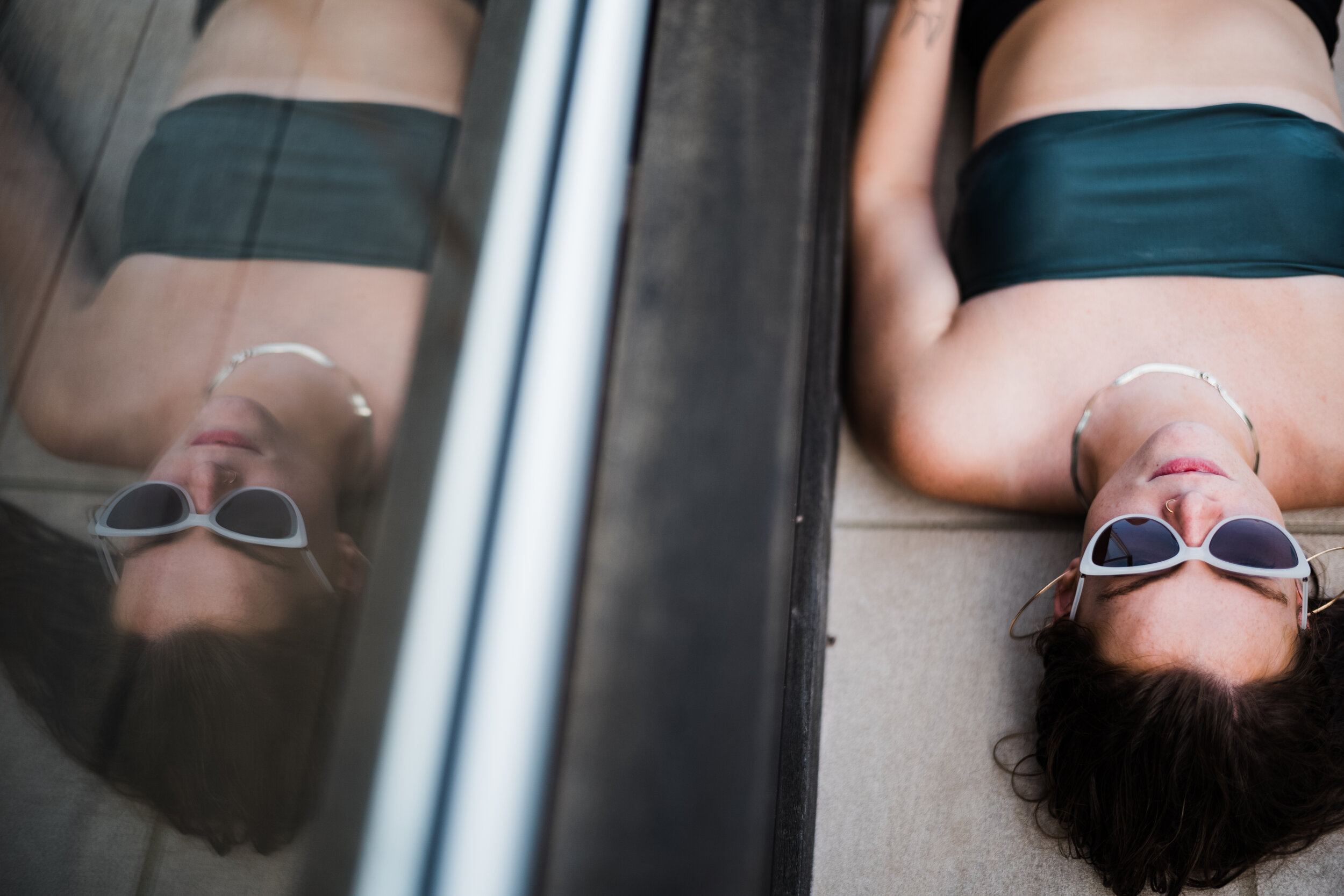 A woman lying on the ground near a building's glass window, wearing sunglasses, a black strapless top, and a choker necklace, with her reflection visible in the window.
