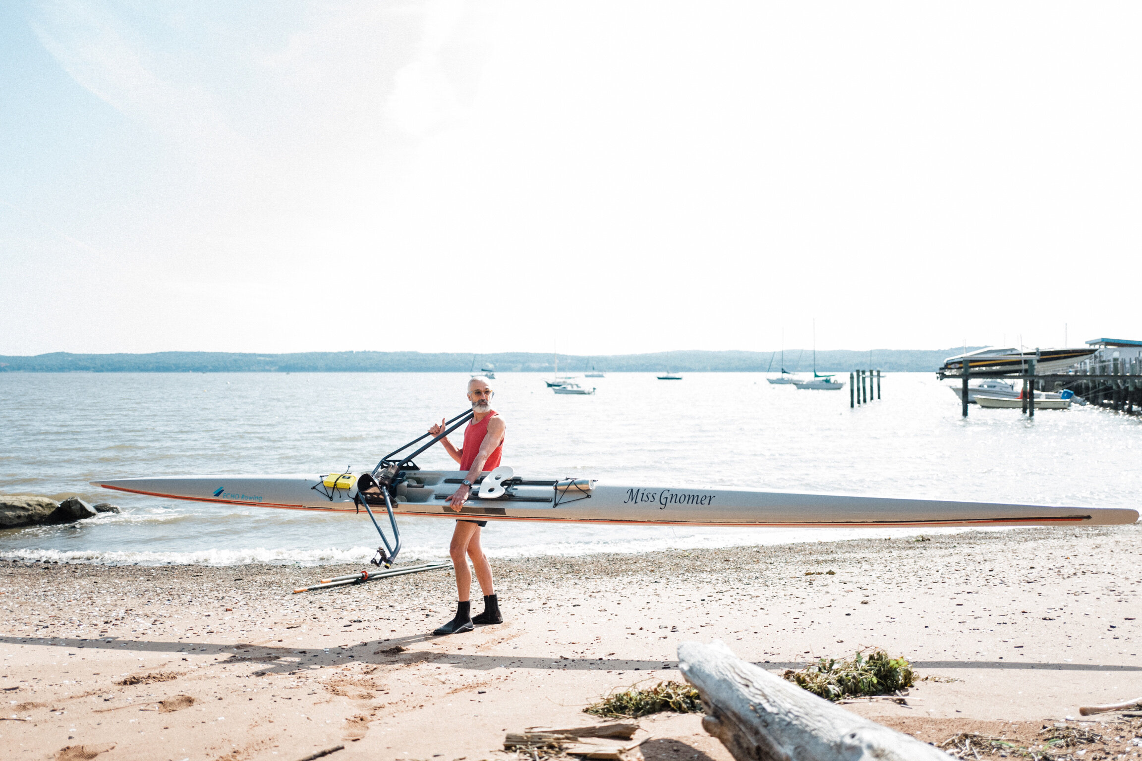 A person holding a racing shell boat on a beach near the water, with boats anchored in the distance and a pier on the right.