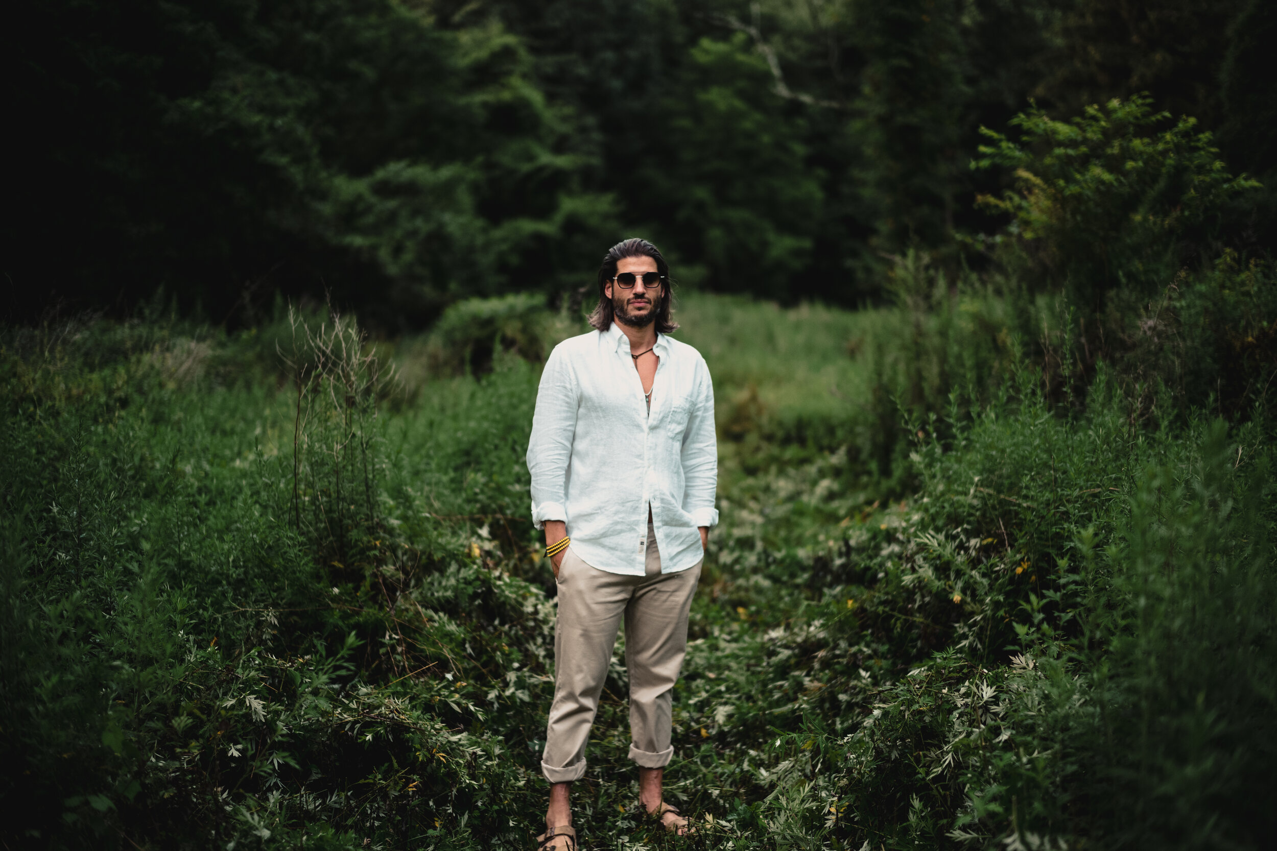 A man with long dark hair, wearing sunglasses, a white shirt, beige pants, and sandals, stands in a lush green outdoor setting with dense foliage and trees in the background.