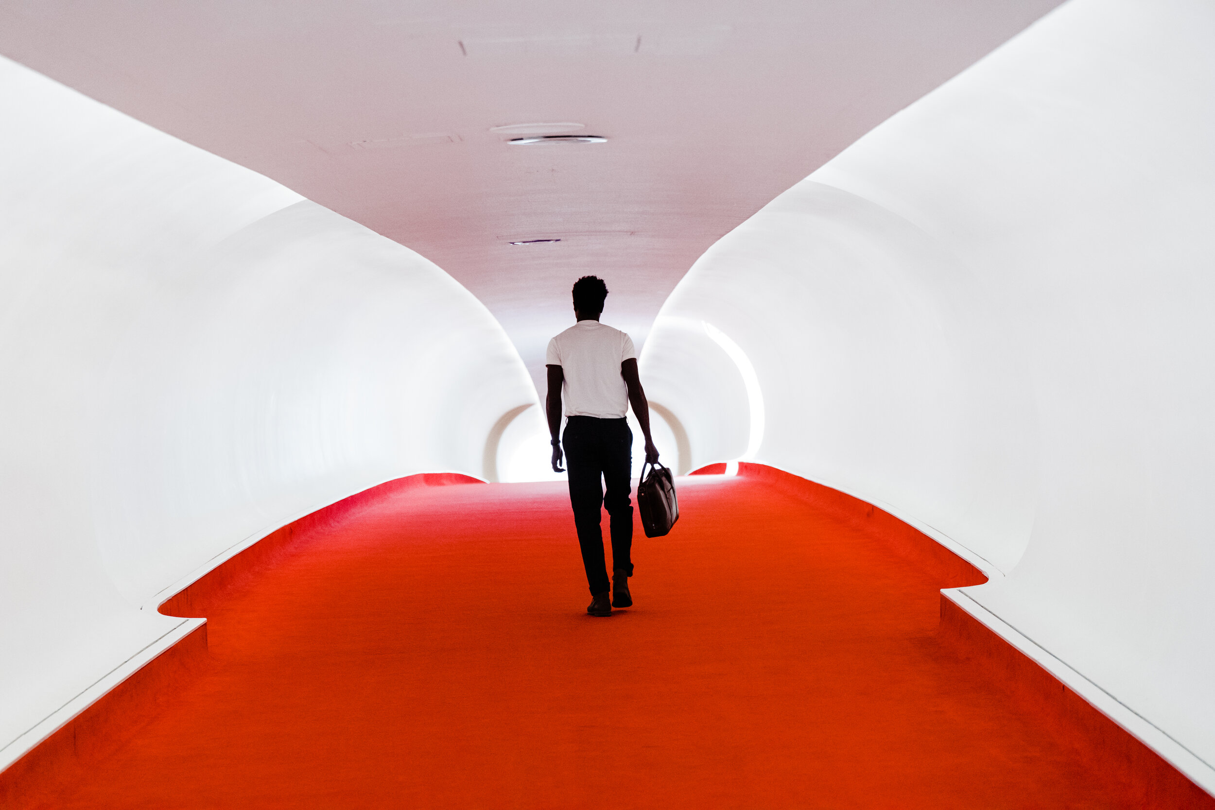 A man walking through a futuristic, white-curved corridor with a red floor, carrying a black bag.