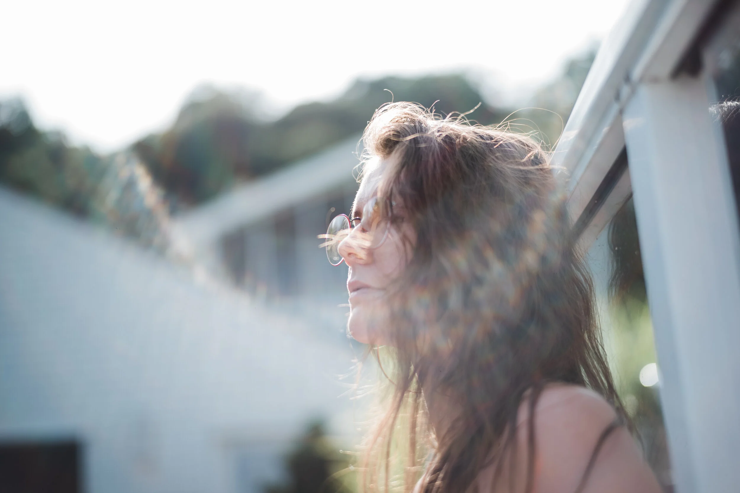 Woman with long curly hair wearing glasses, enjoying sunlight outdoors near a window.