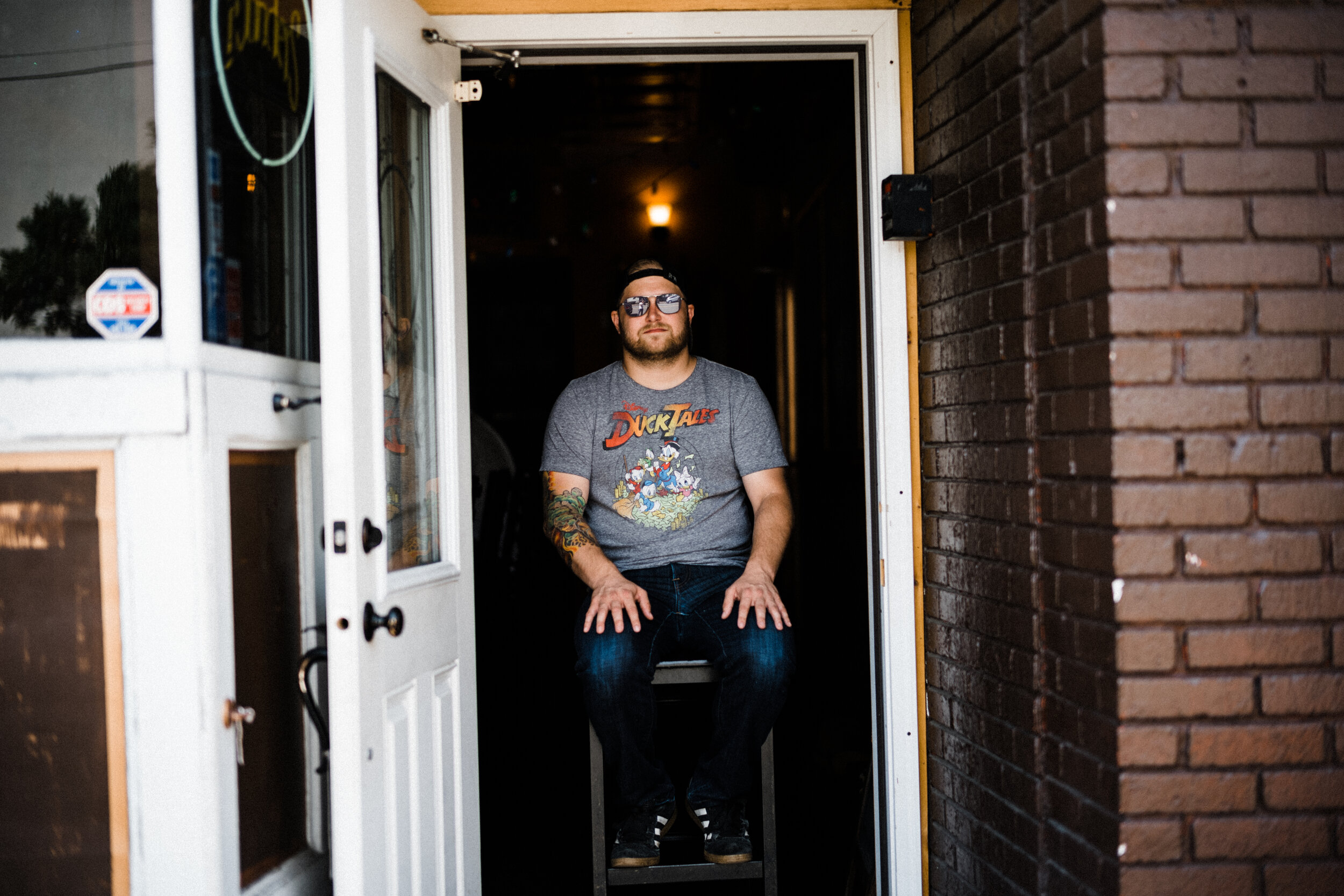 A man with tattoos wearing sunglasses and a DuckTales t-shirt sits on a toilet inside a restroom, partially visible through an open door.