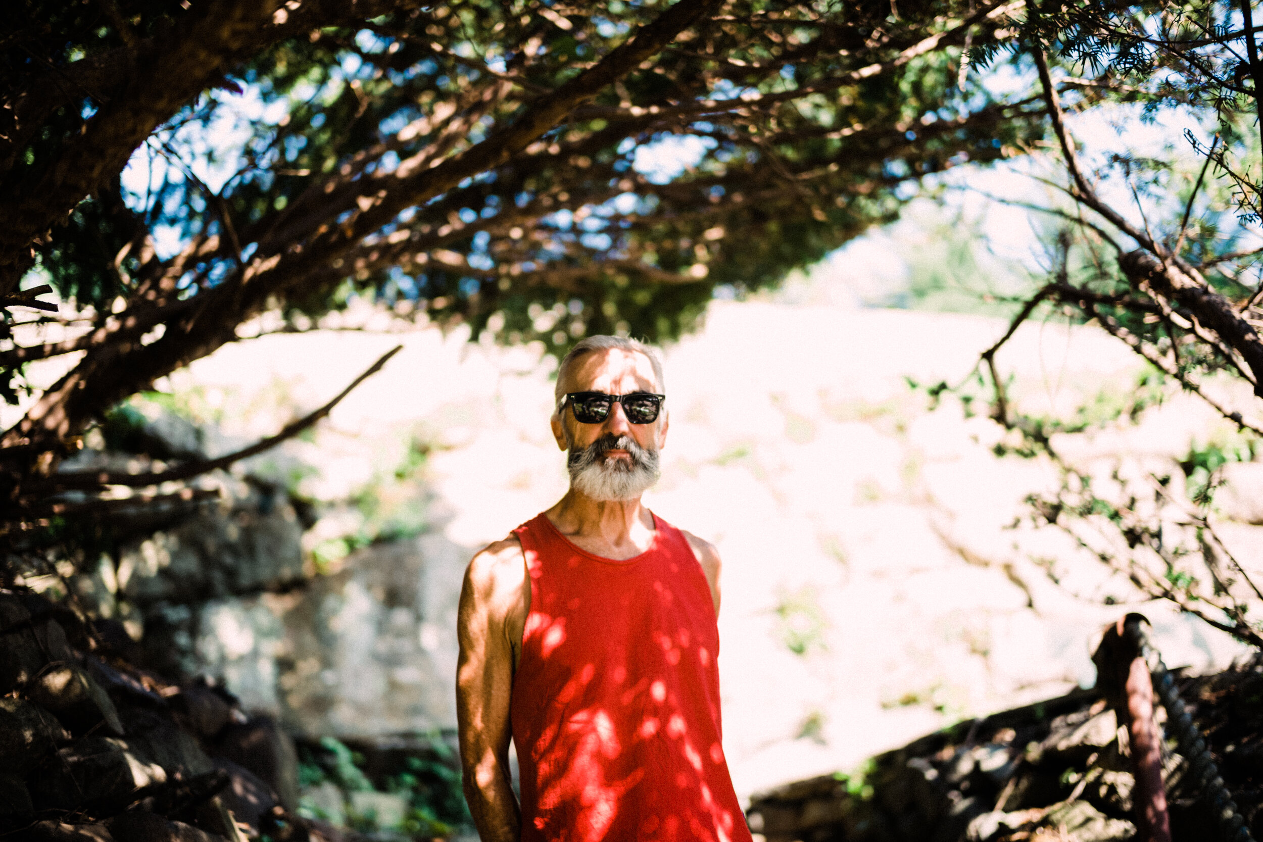 An older man with a gray beard and mustache wearing sunglasses and a red sleeveless shirt standing outdoors, shaded by tree branches.