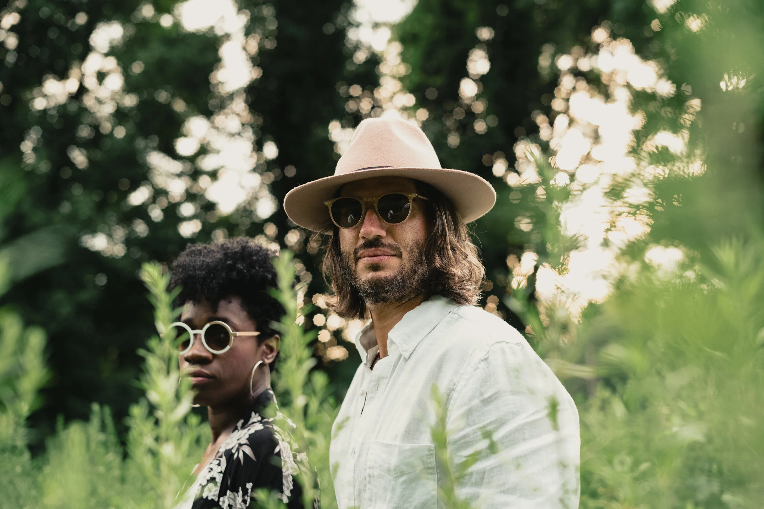 Two people standing among greenery outdoors, wearing sunglasses and hats, with trees and sunlight in the background.