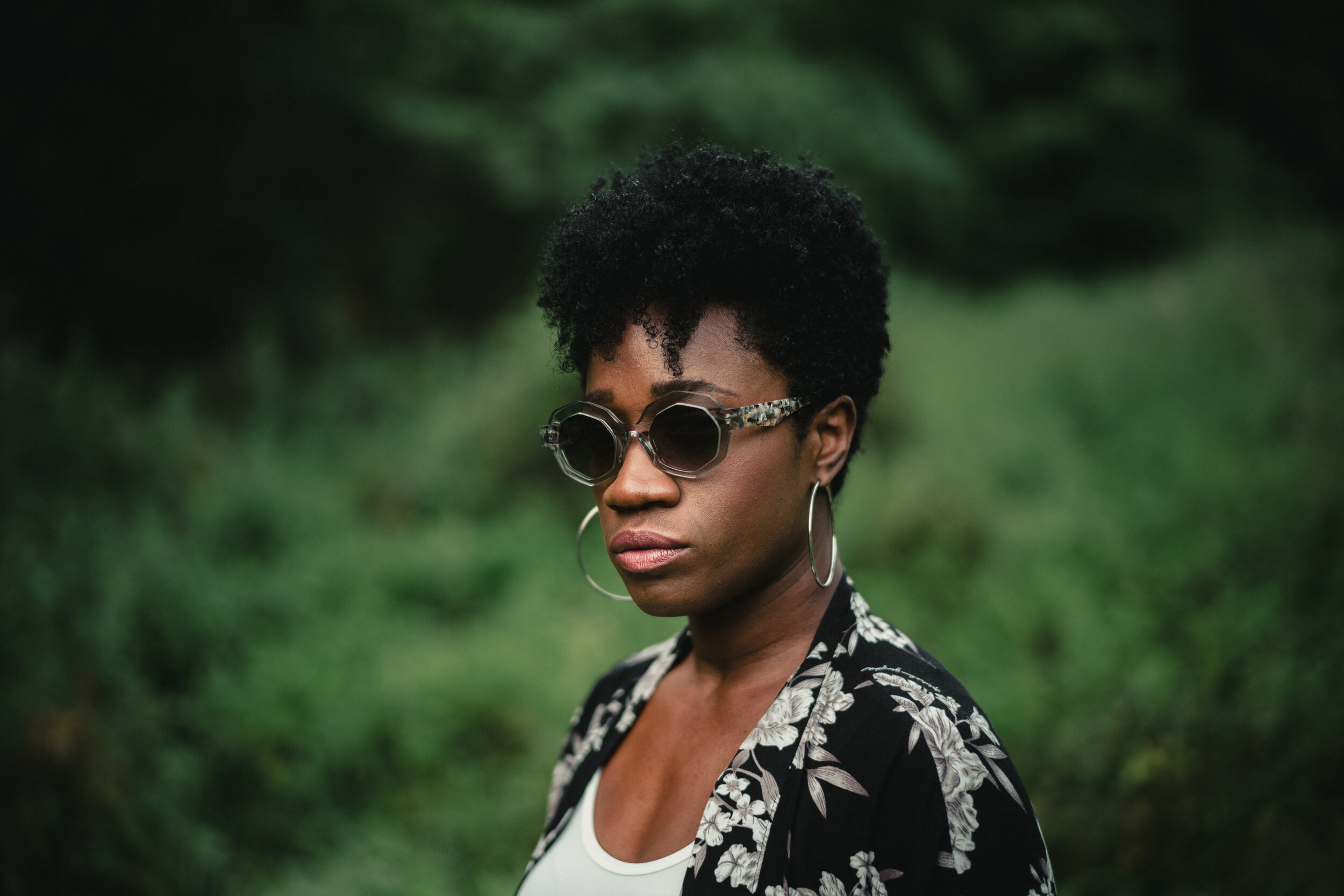 A woman with dark skin and short, curly black hair wearing large sunglasses, hoop earrings, a floral patterned jacket, and a white shirt, standing outdoors with green foliage in the background.