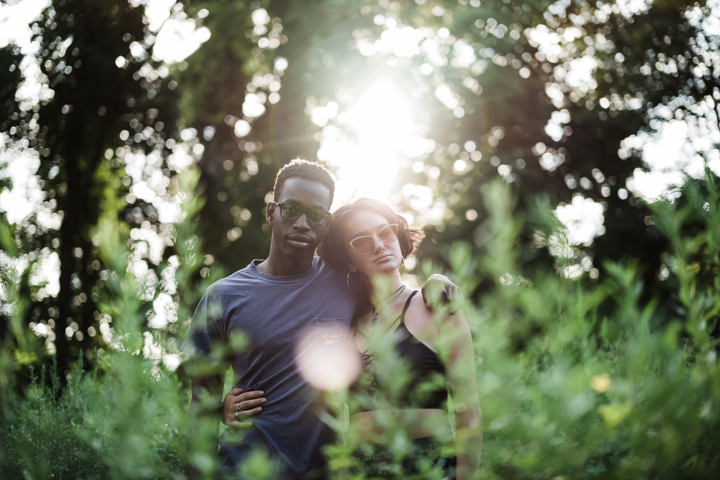 Two young women standing close together outdoors, with sunlight shining through trees in the background. One woman has short hair, wears glasses, and a casual t-shirt. The other woman has longer hair, wears sunglasses and a black top. They are in a n