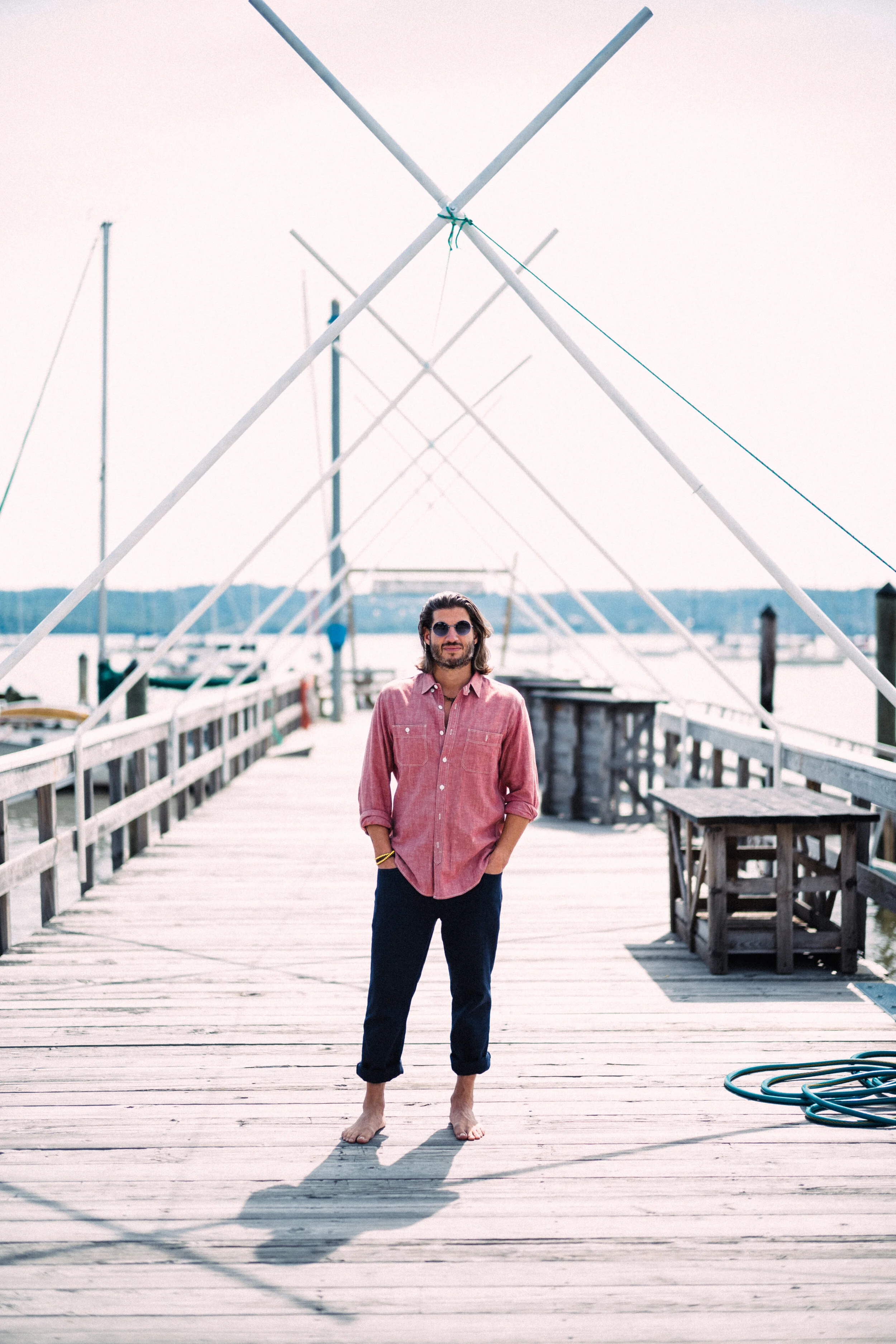 A man with long hair, sunglasses, a red button-up shirt, and rolled-up dark pants standing barefoot on a wooden dock near a body of water with sailboat masts in the background.