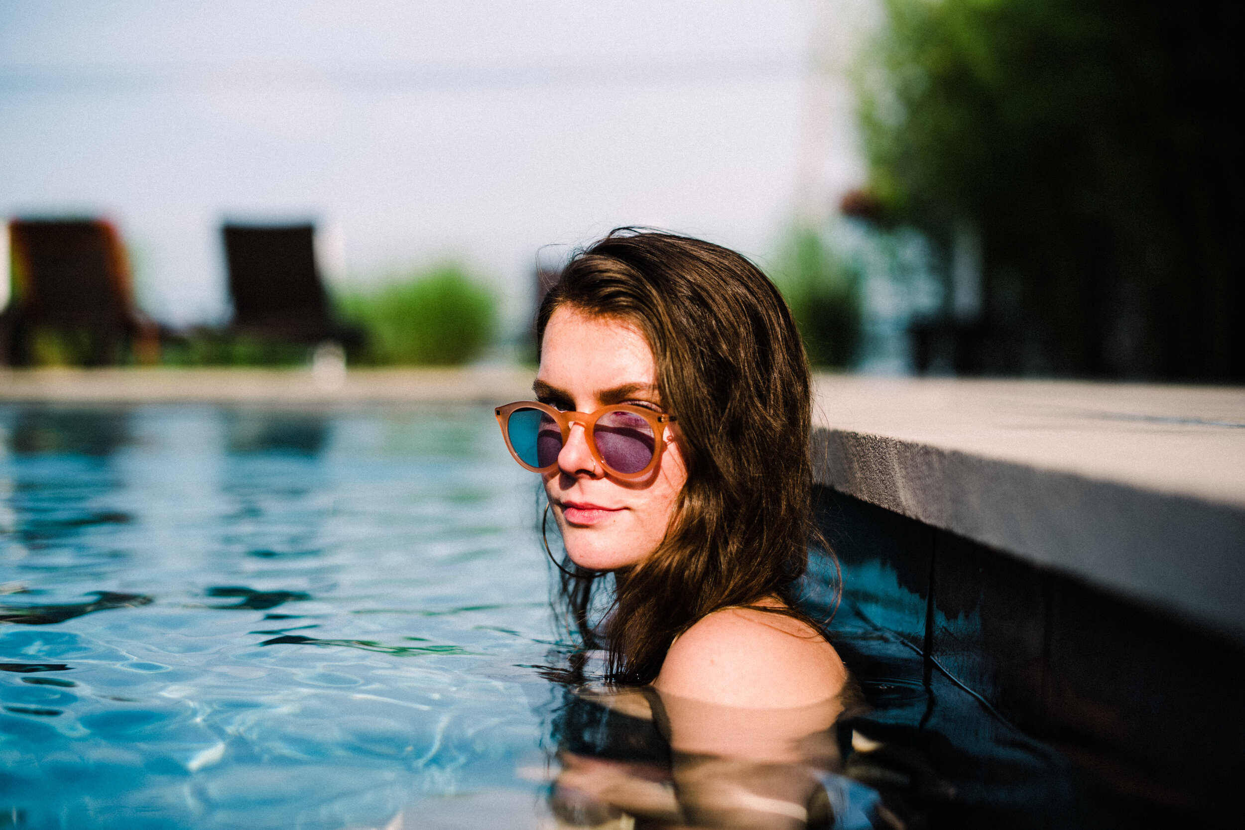 Woman with brown hair wearing sunglasses relaxing in a swimming pool near the edge on a sunny day.