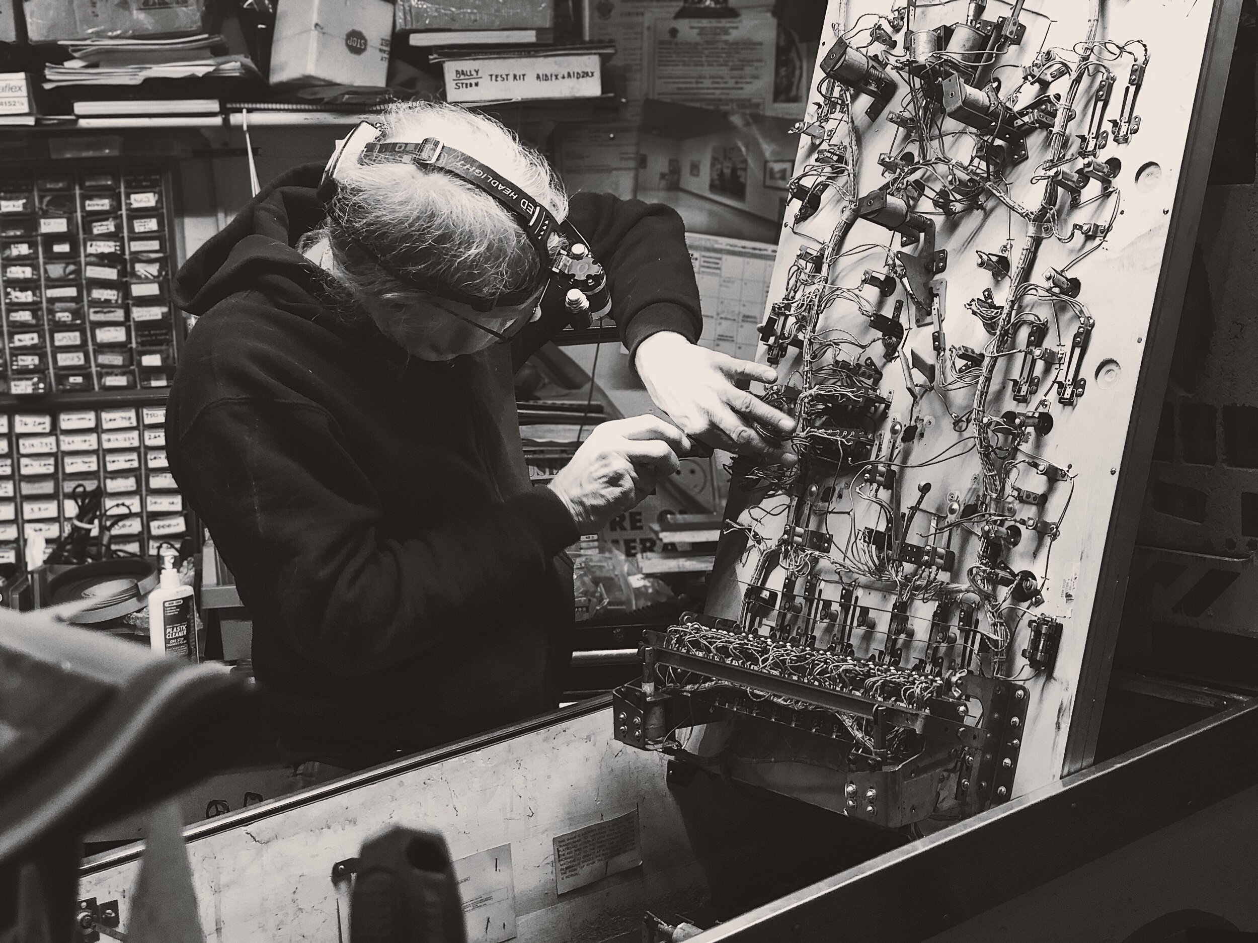A person wearing a hooded sweatshirt and a headlamp working on an electronic circuit board with wires and components in a workshop