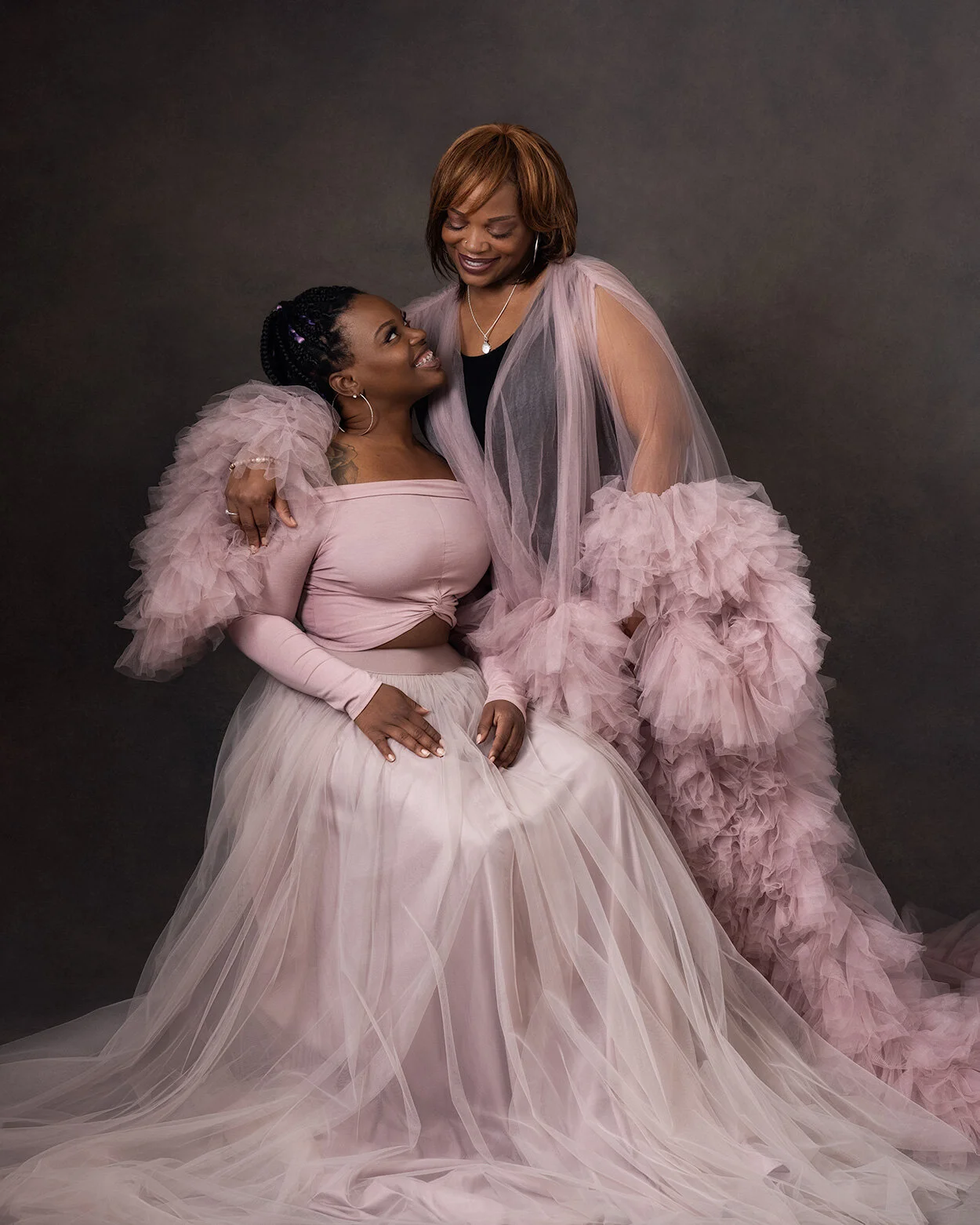 Two women dressed in pink and blush tulle dresses sharing a joyful moment against a dark gray background. One woman is seated, the other standing, with the standing woman's arm around the seated woman's shoulder.