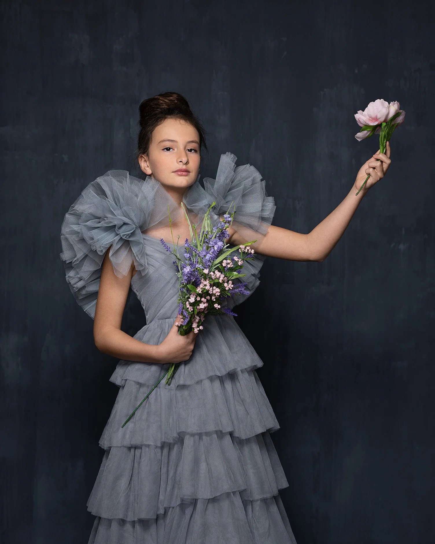 A young girl wearing a ruffled, gray dress with puffed sleeves, holding a bouquet of purple and pink flowers, standing against a dark backdrop.