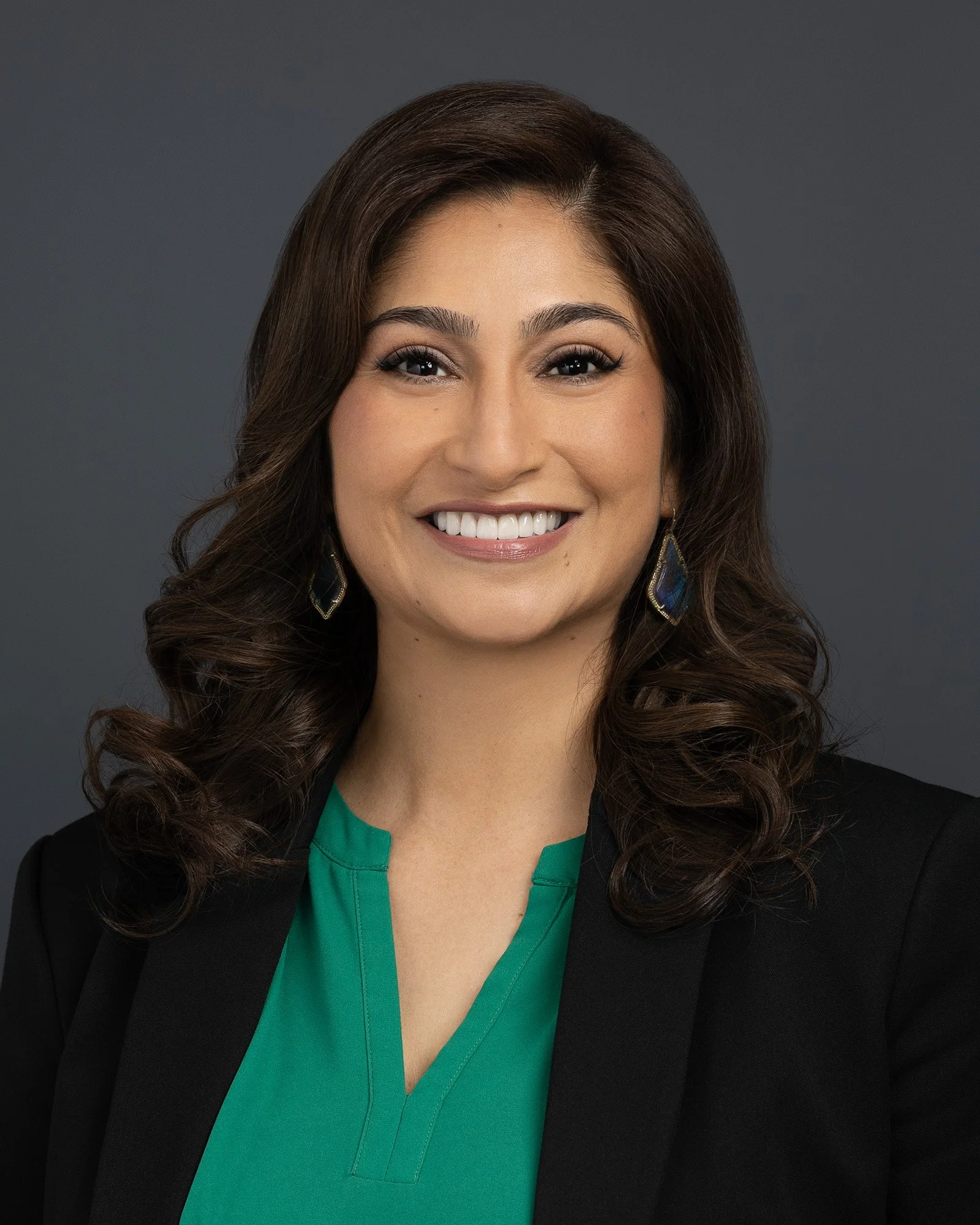 Headshot of a woman with wavy brown hair, wearing a black blazer and green top, smiling, against a dark gray background.