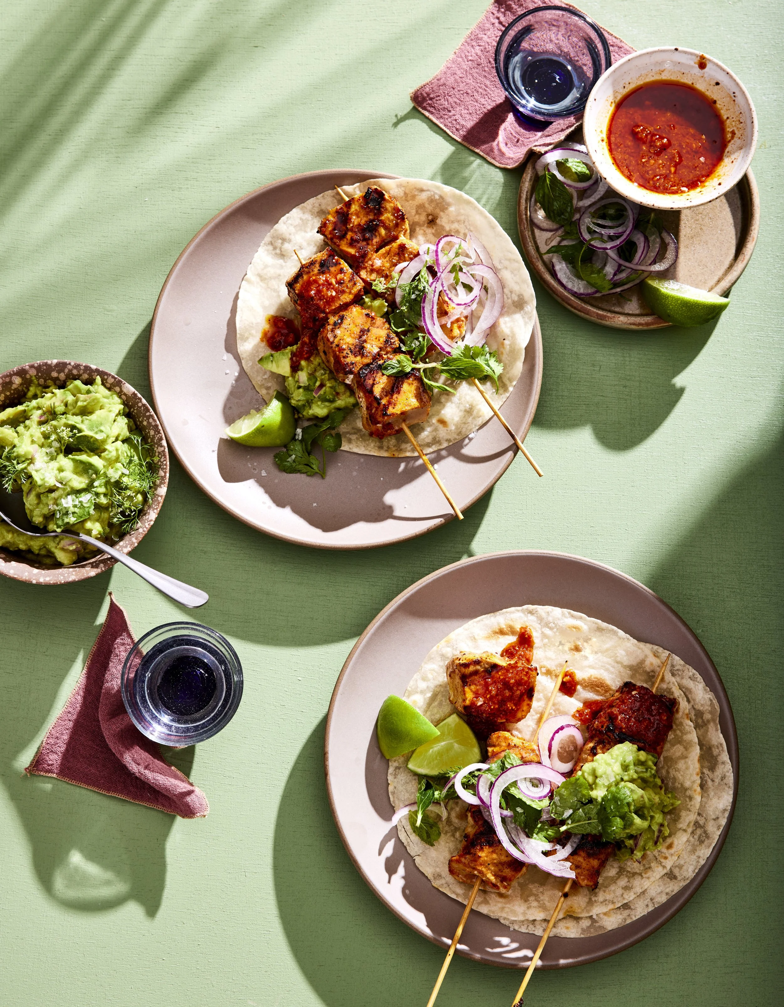 Plate with skewered grilled chicken, lettuce, sliced red onions, and cilantro, served on a soft taco. Also on the table are a bowl of guacamole, a bowl of red salsa with lime wedges, and a glass of water, all arranged on a green table with pink and red napkins.