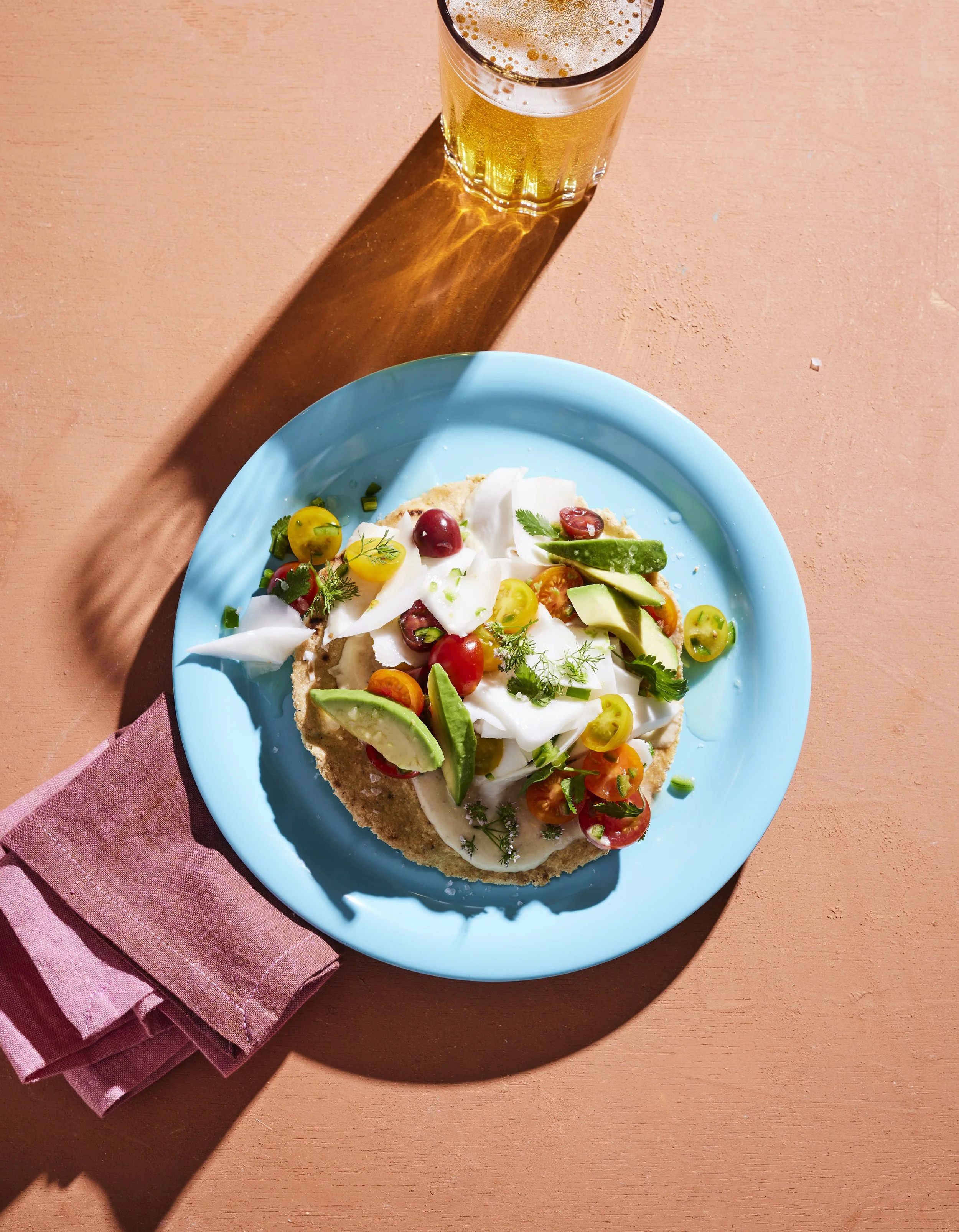 A slice of avocado and tomato pizza topped with fresh herbs on a blue plate, with a glass of beer nearby, on a pink placemat and table.