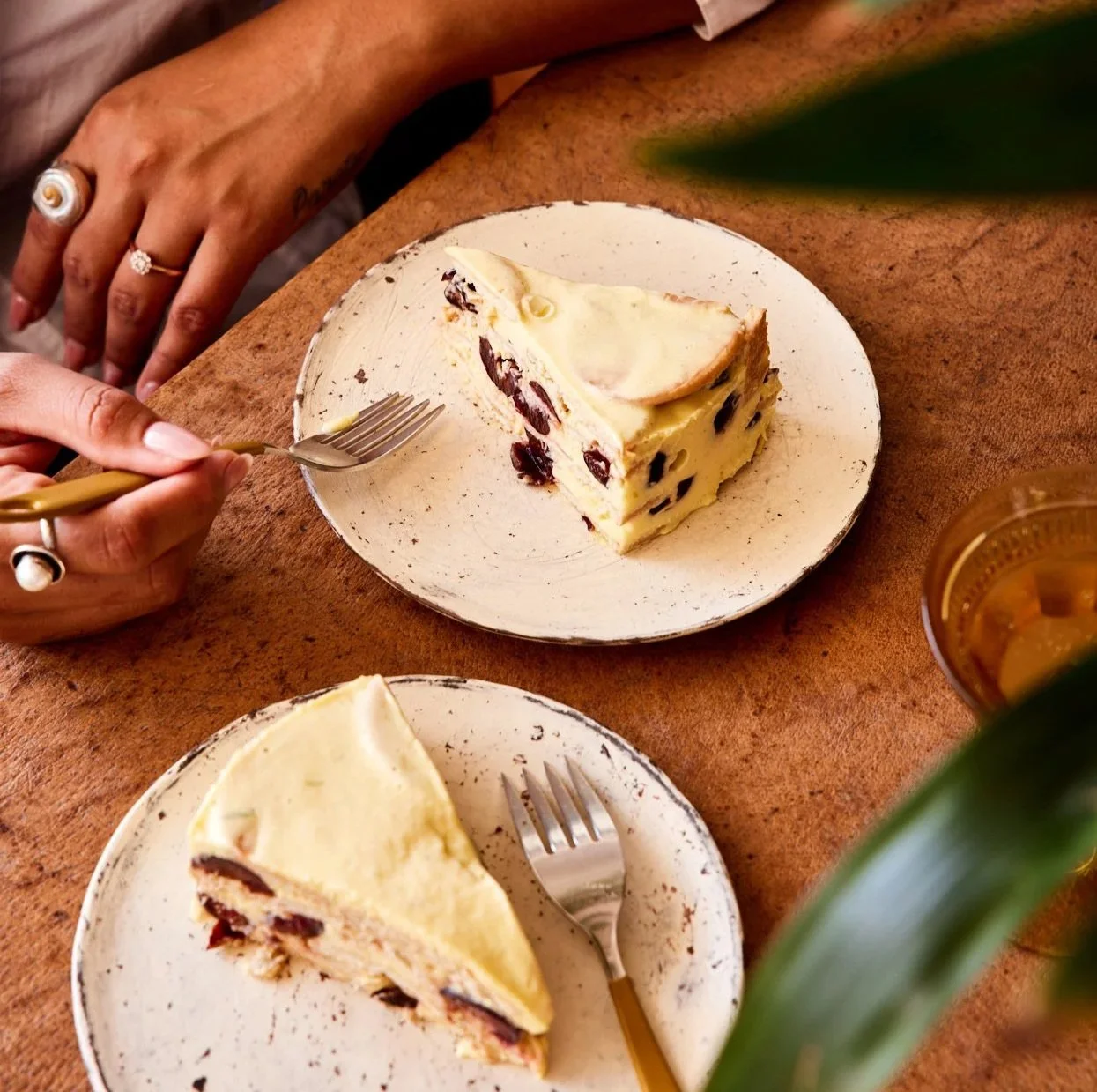 Slice of cherry cheesecake on a white plate with a fork, placed on a wooden table with a dollar drink in a clear cup and a woman's hand wearing rings in the background.