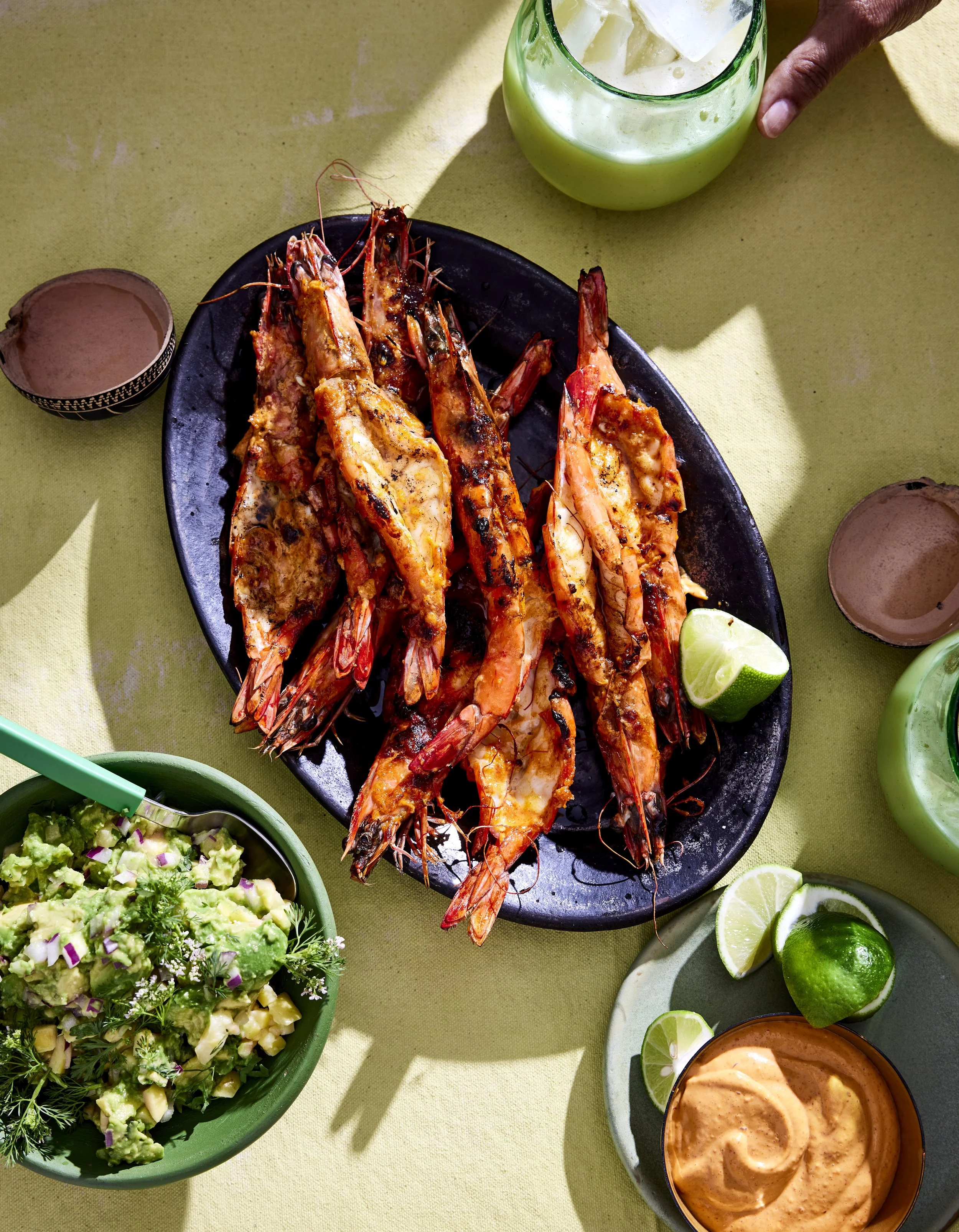 A platter of grilled shrimp with lemon halves, a bowl of guacamole, and bowls of sauces and dips on a yellow tablecloth.