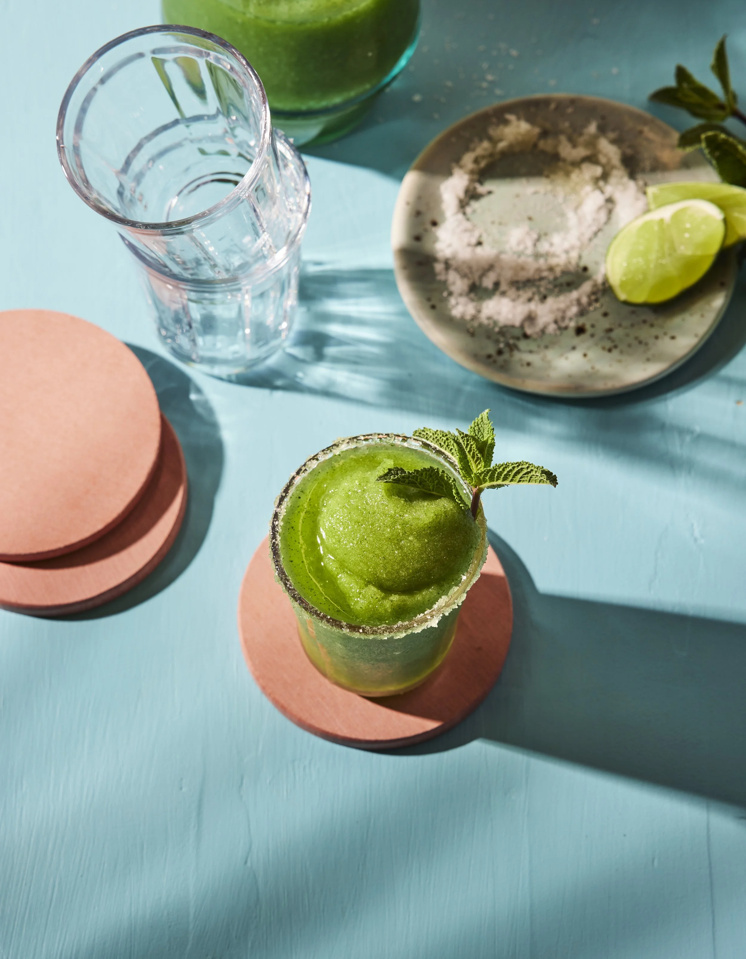Top view of a vibrant green cocktail garnished with mint leaves in a salt-rimmed glass on a turquoise surface, with a lime wedge on a plate nearby and a stack of pink coasters.
