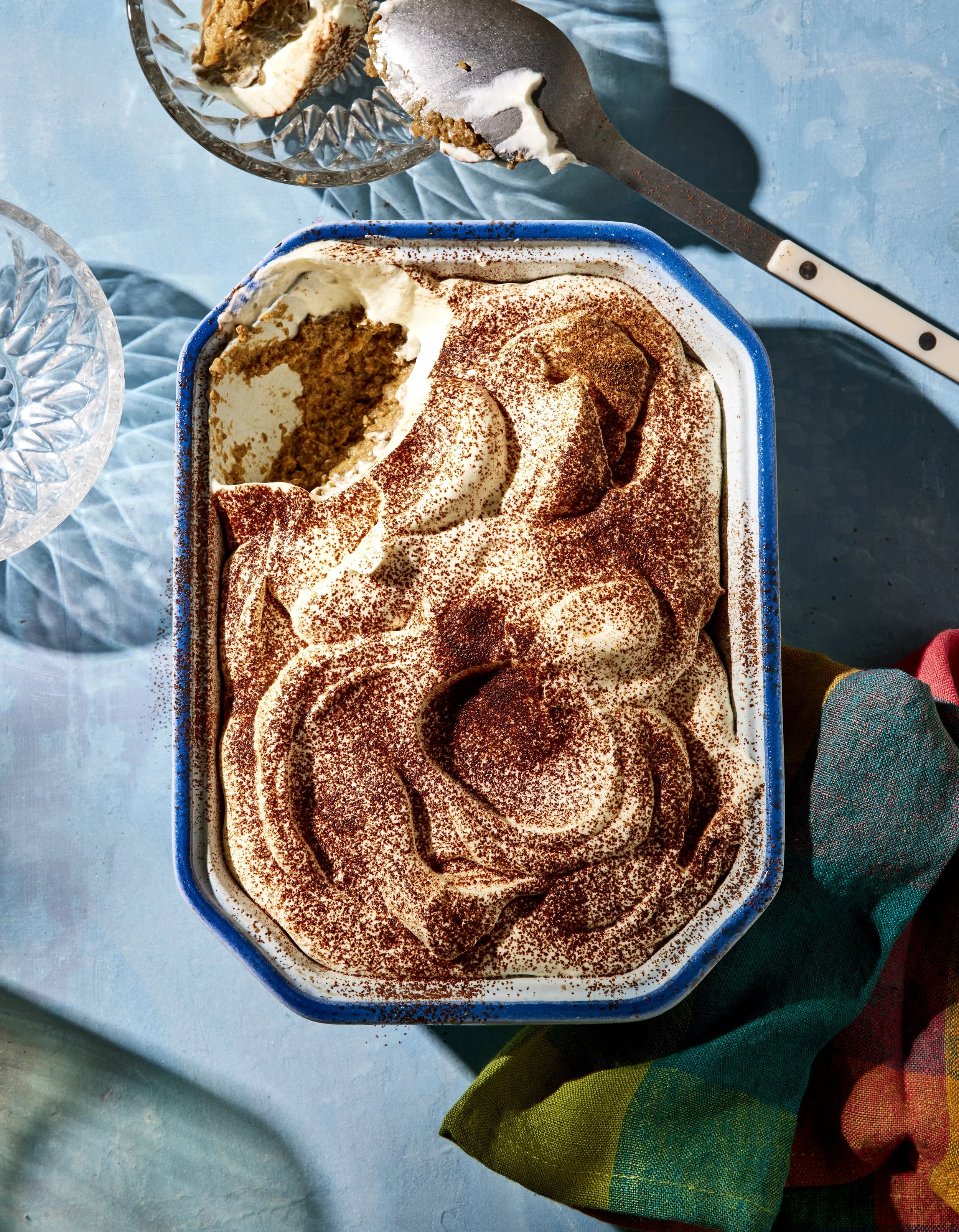A rectangular ceramic dish filled with tiramisu topped with cocoa powder. A scoop is missing, revealing the layered cake inside. The dish is placed on a light blue surface, with a colorful cloth partially visible at the bottom right corner.