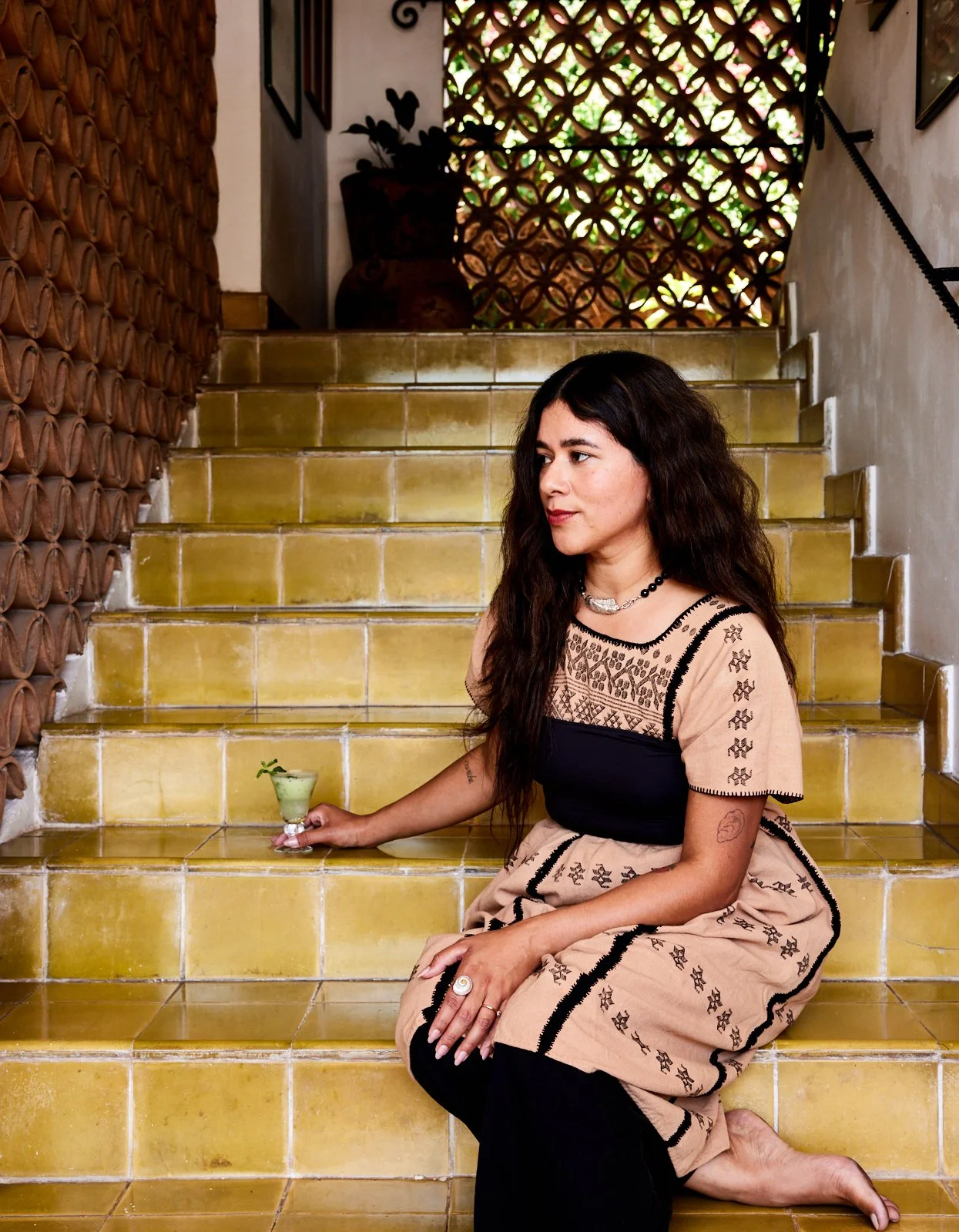 A woman with long, wavy dark hair sitting on yellow tiled stairs in a rustic, tropical-style setting. She is wearing a beige dress with black embroidered accents and black leggings. She is holding a small glass of green drink garnished with a lime wedge and mint. The background features decorative wooden lattice and potted plants, with sunlight filtering through.