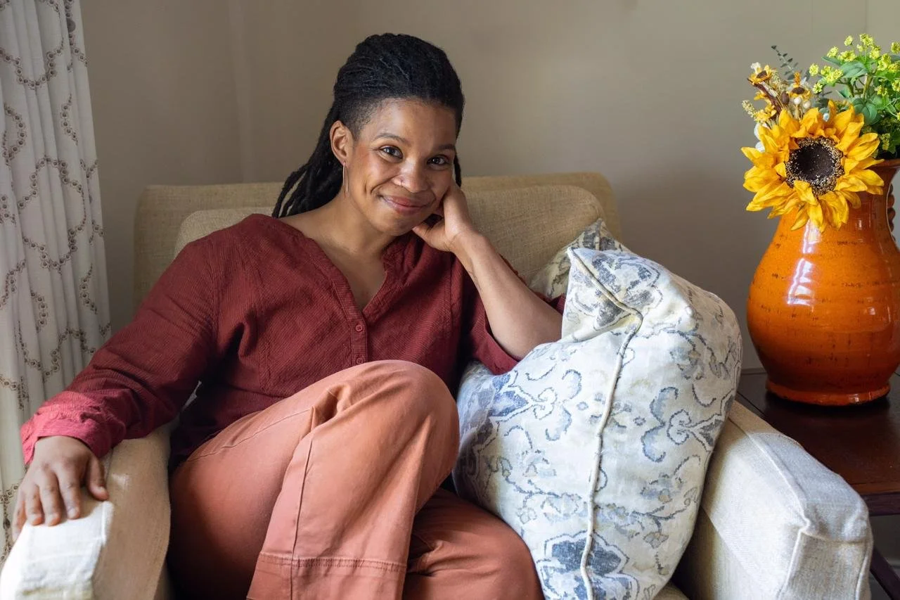 Brown skinned woman with long dreadlocks sits in a chair with one knee up, smiling at the camera