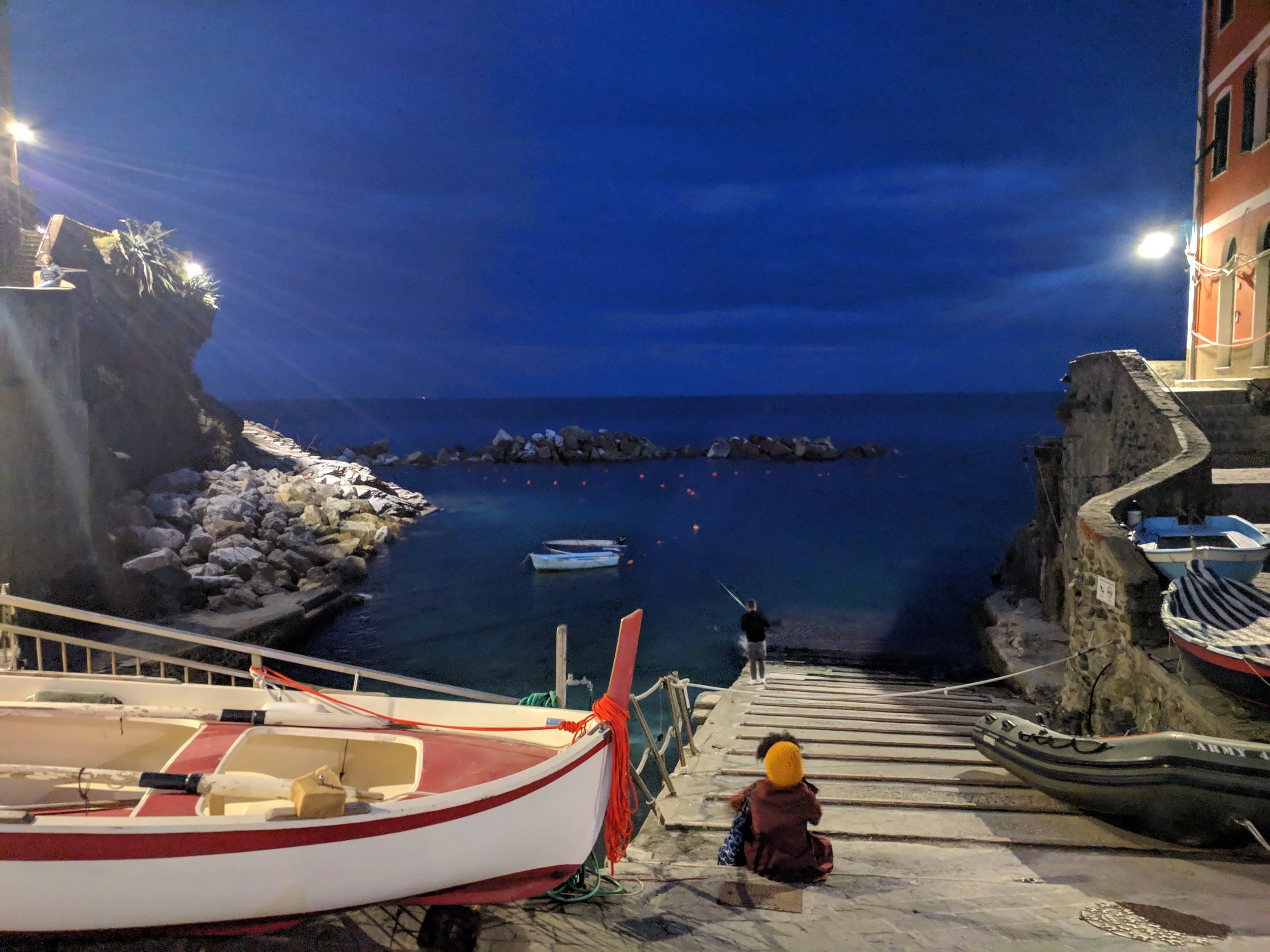 View of the sea. A person in a burgundy jacket and mustard hat sits back to us; it's shortly after sunset.