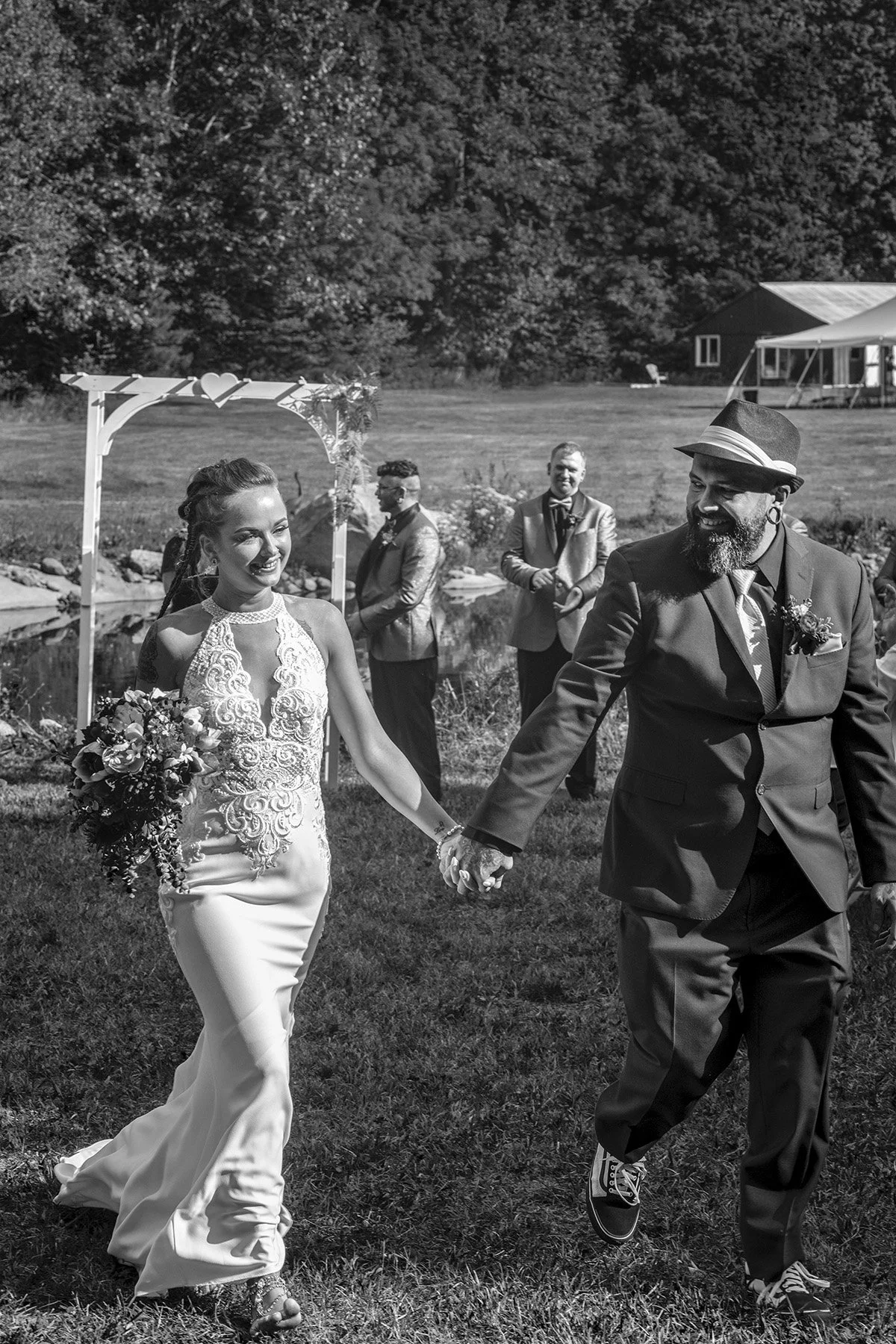 A bride and groom holding hands and smiling, walking outdoors at their wedding, with bridesmaids and groomsmen in the background near a pond and a decorative arch.
