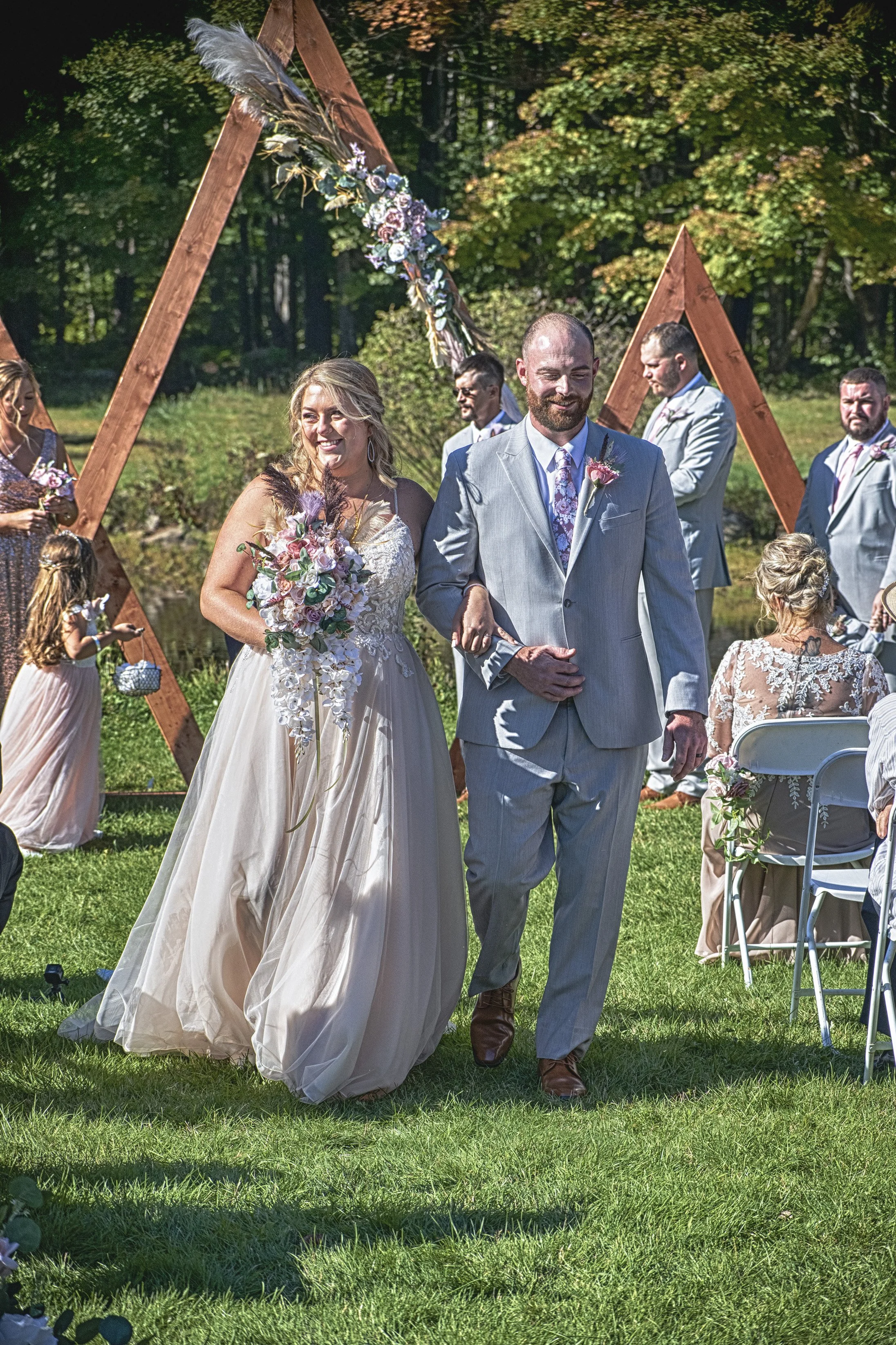 Bride and groom walking down the aisle during their outdoor wedding ceremony, decorated with floral arrangements and wooden triangular arch, surrounded by friends and family.