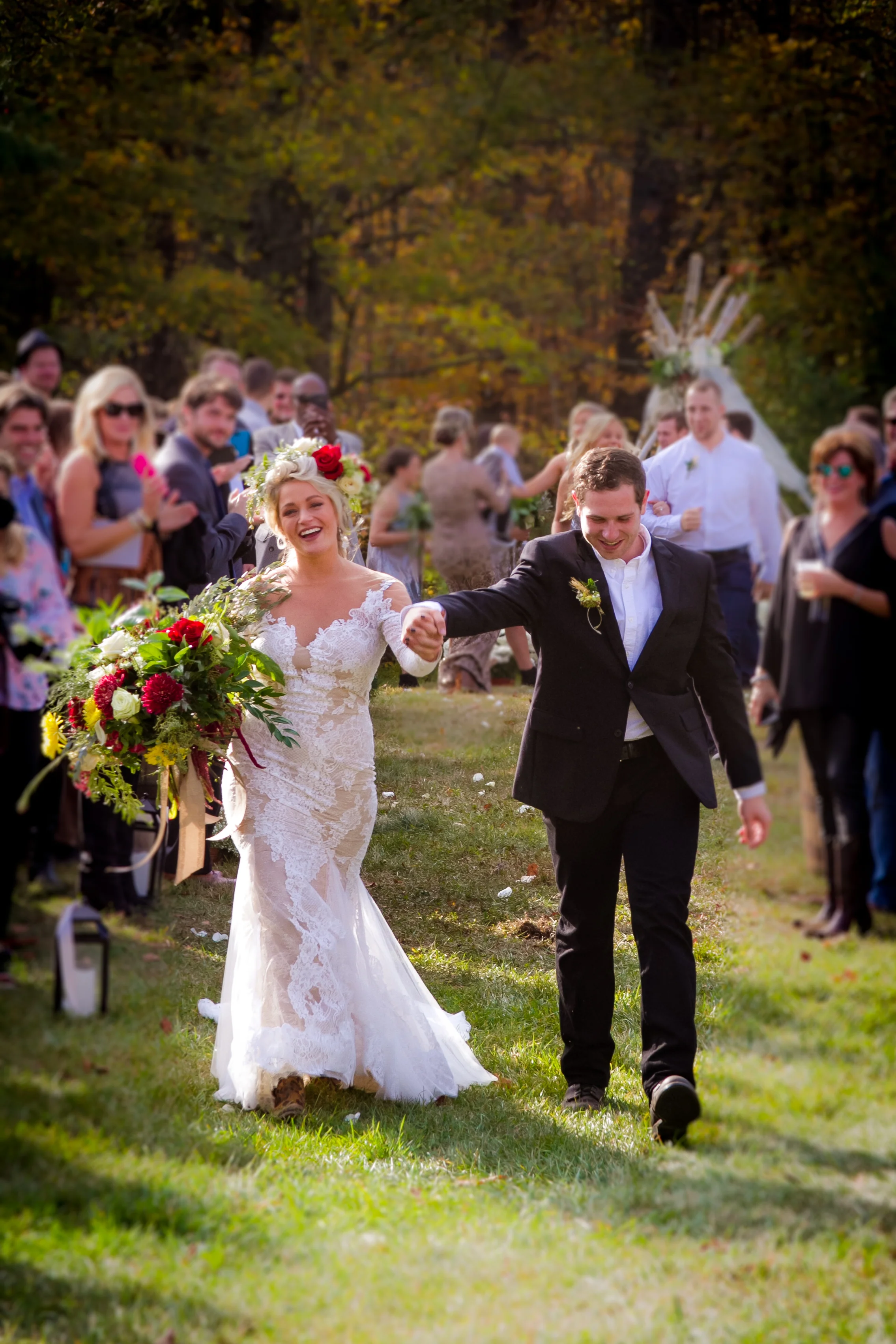 Here, Lexee and Brandon stroll back down the aisle after saying "I do". Lexee is wearing a long sleeved mermaid lace gown. Her stunning bouquet contains lovely fall colors such as red, white, yellow and green match the flower crown atop her head. Brandon is donning a simple black suit, with no tie, noting the casual and comfortable feel of their wedding. In the background, you can see the teepee that they got married in front of.
