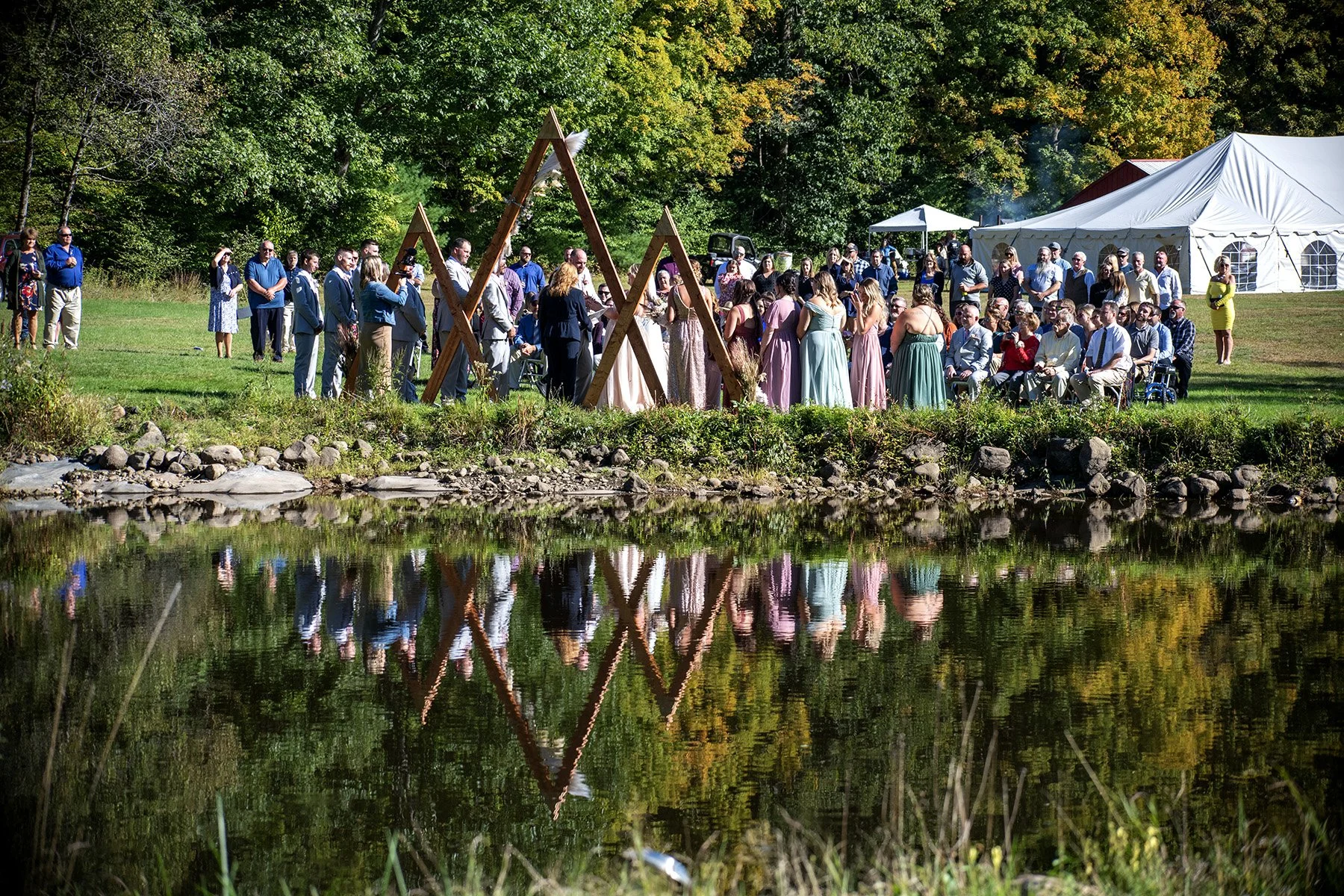 A large gathering of people at an outdoor wedding ceremony near a pond, with trees and a white tent in the background. The reflection of the scene is visible in the water.