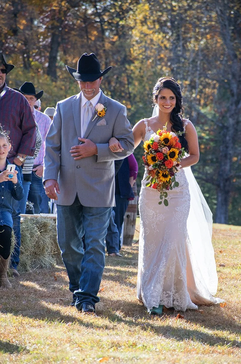 A bride walking down the aisle with a man, likely her father, during an outdoor wedding. The bride wears a lace wedding dress and holds a bouquet of sunflowers, roses, and greenery. The man wears a light gray suit, a white shirt, a light-colored tie,