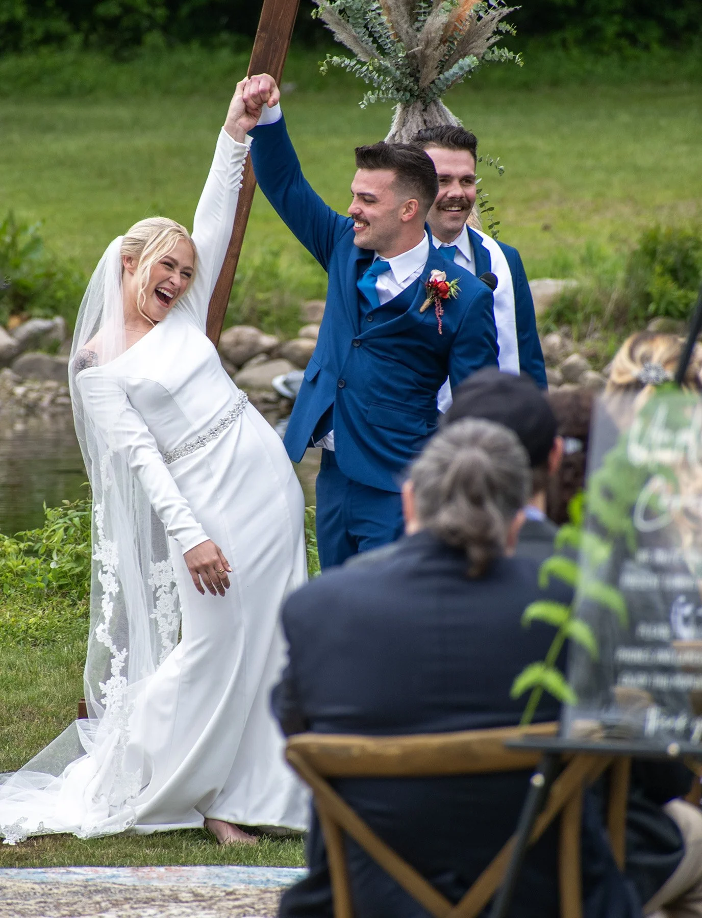 A bride and groom celebrate at their outdoor wedding ceremony, holding hands and smiling, with a priest or officiant in the background near a decorative plant. Guests are seated in front.