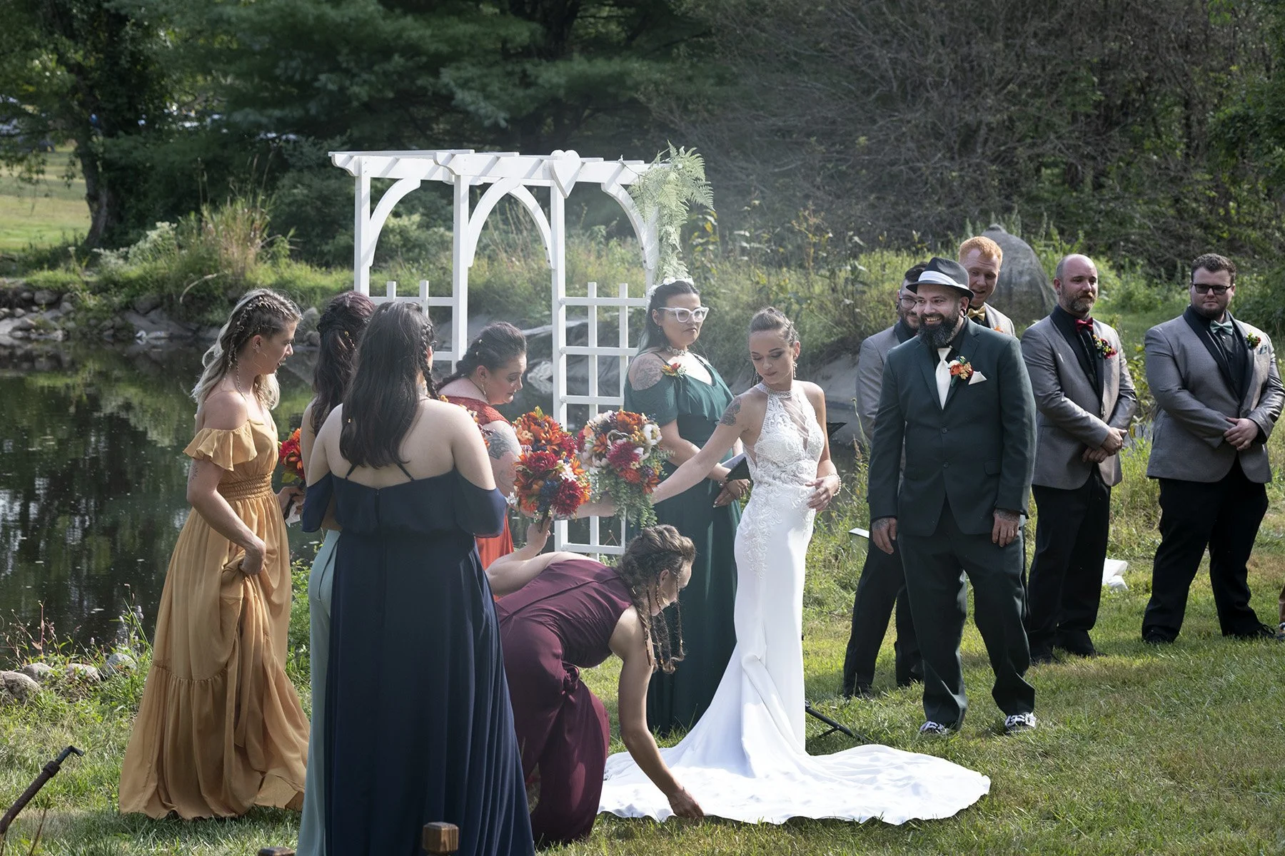 A wedding ceremony outdoors by a pond, with a bride in a white wedding dress holding a bouquet and a groom in a dark suit and hat, surrounded by bridesmaids in colorful dresses and groomsmen in suits, with a white arbor decorated with greenery in the