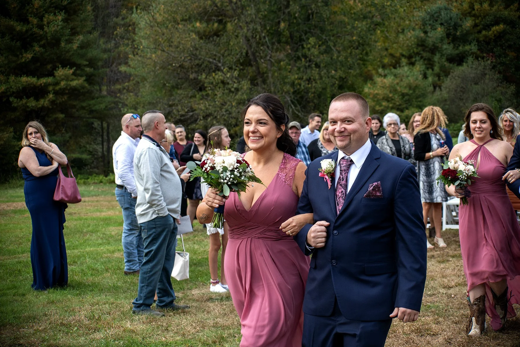 A wedding procession outdoors with a smiling woman in a pink dress holding a bouquet and a man in a navy suit. Other guests in formal attire hold bouquets and stand on a grassy area with trees in the background.