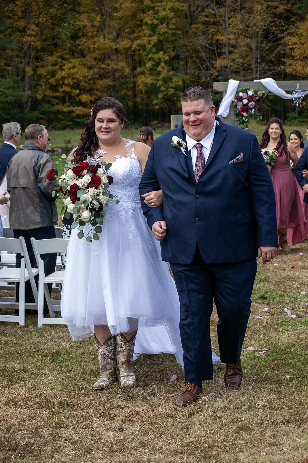 Bride and groom walking down the aisle at an outdoor wedding ceremony, with guests seated on white chairs and a decorated arch in the background, surrounded by fall foliage.