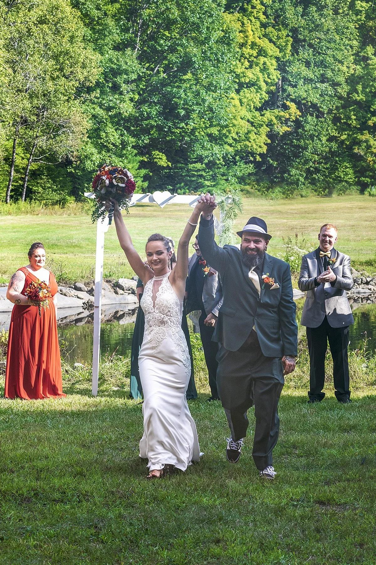 A newlywed couple walking down the aisle outdoors, holding hands and raising a bouquet, with friends in the background celebrating.