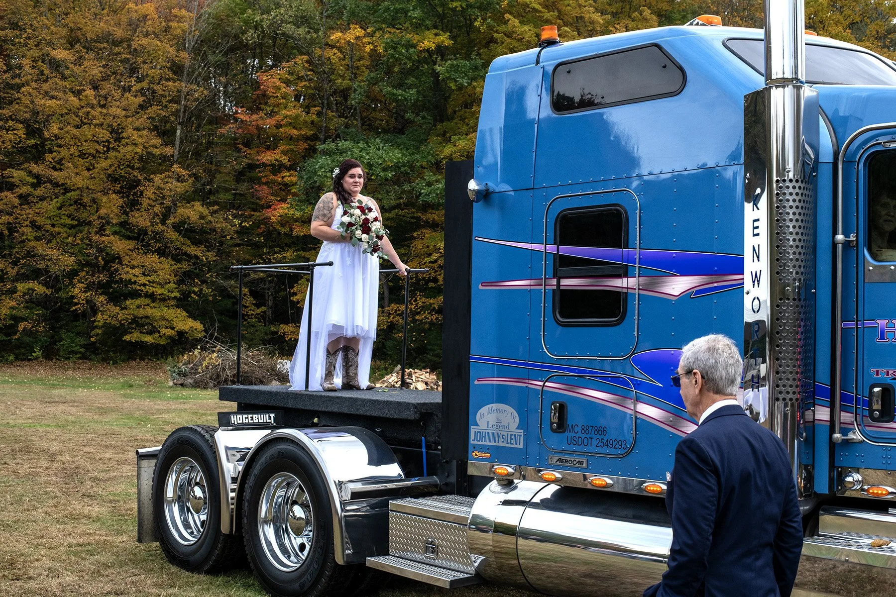 A woman in a white dress with tattoos and cowboy boots stands on a mobile stage holding a bouquet of flowers, with a colorful background of autumn trees. A man in a suit is in the foreground, looking towards her.