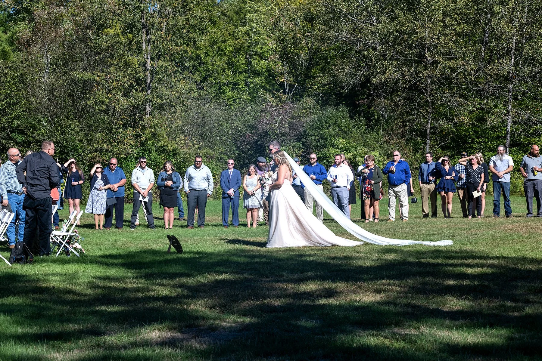 Wedding ceremony outdoors with bride and groom standing in front of a group of guests, surrounded by trees and grass on a sunny day.