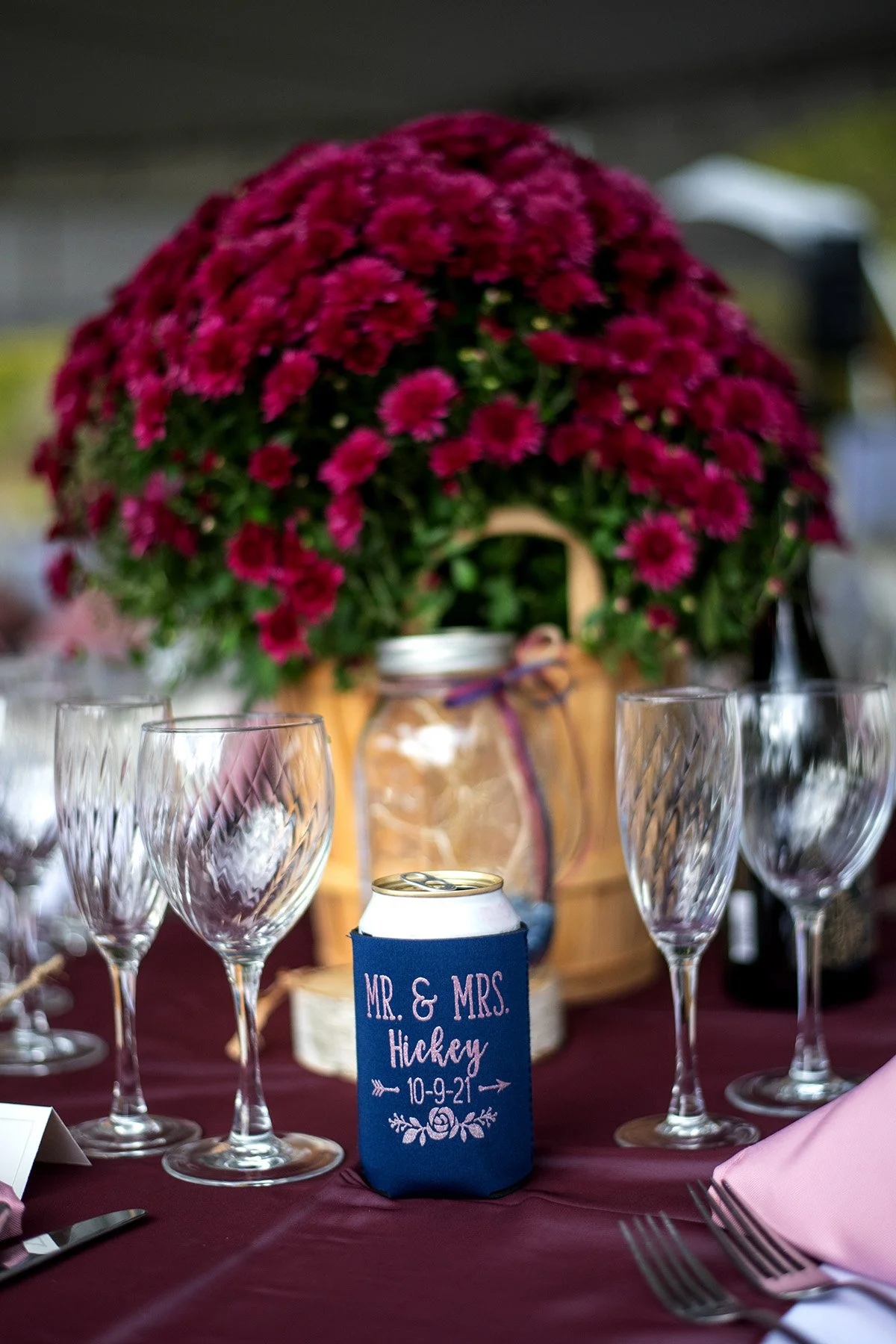 A wedding table setting with a large bouquet of pink flowers in a basket, several empty wine glasses, a can of soda in a blue koozie engraved with "MR. & MRS. Hickey 10-9-21," and pink napkins on a burgundy tablecloth.