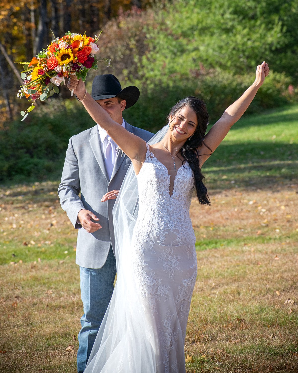 A woman in a white wedding dress holding a bouquet of colorful flowers, smiling with her arms raised, outdoors in a park-like setting with green trees and grass, with a man in a gray suit and black cowboy hat behind her.
