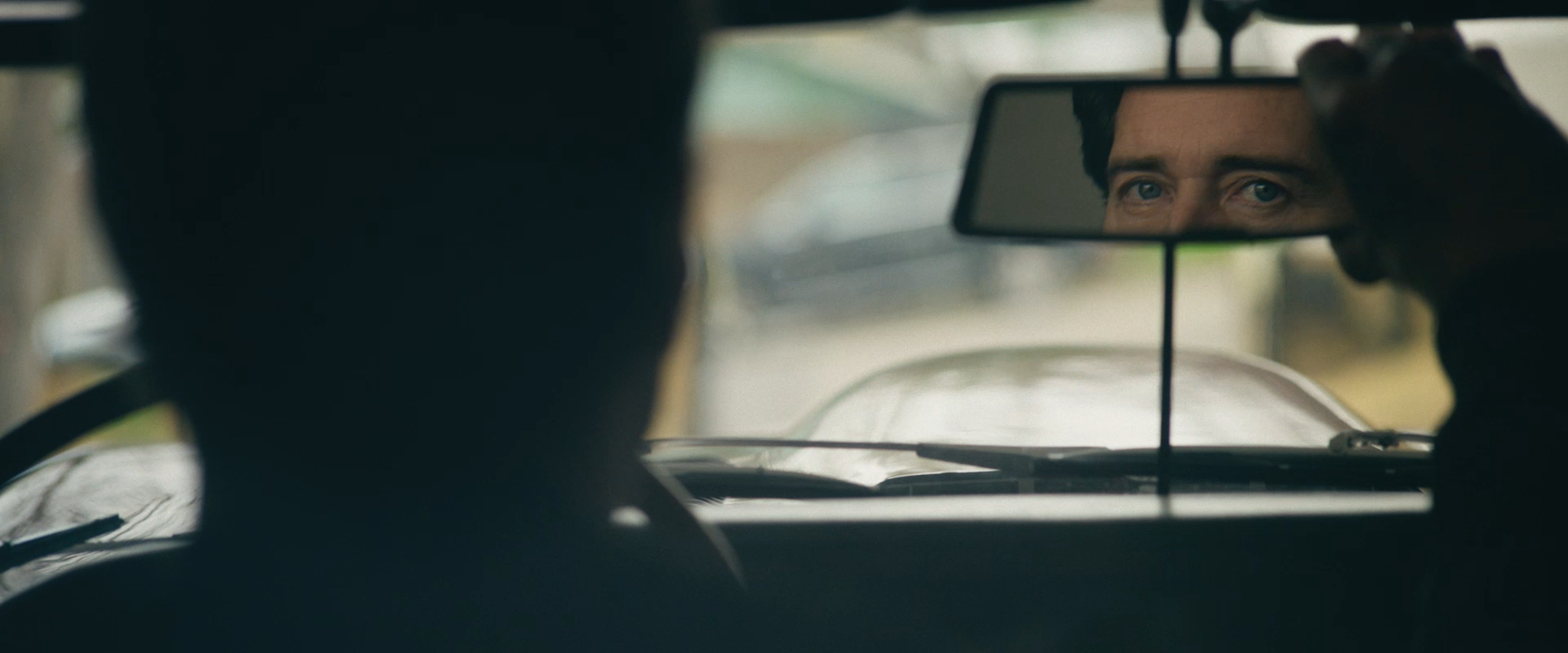 A man is adjusting his rearview mirror and looking at his reflection with blue eyes while seated inside a car.