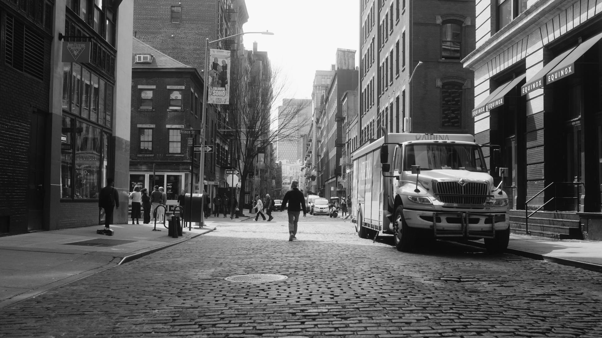 A black and white photo of a city street with pedestrians and a delivery truck parked on the cobblestone road. Multi-story buildings line the street, and there are signs, parked cars, and a tree in the scene.