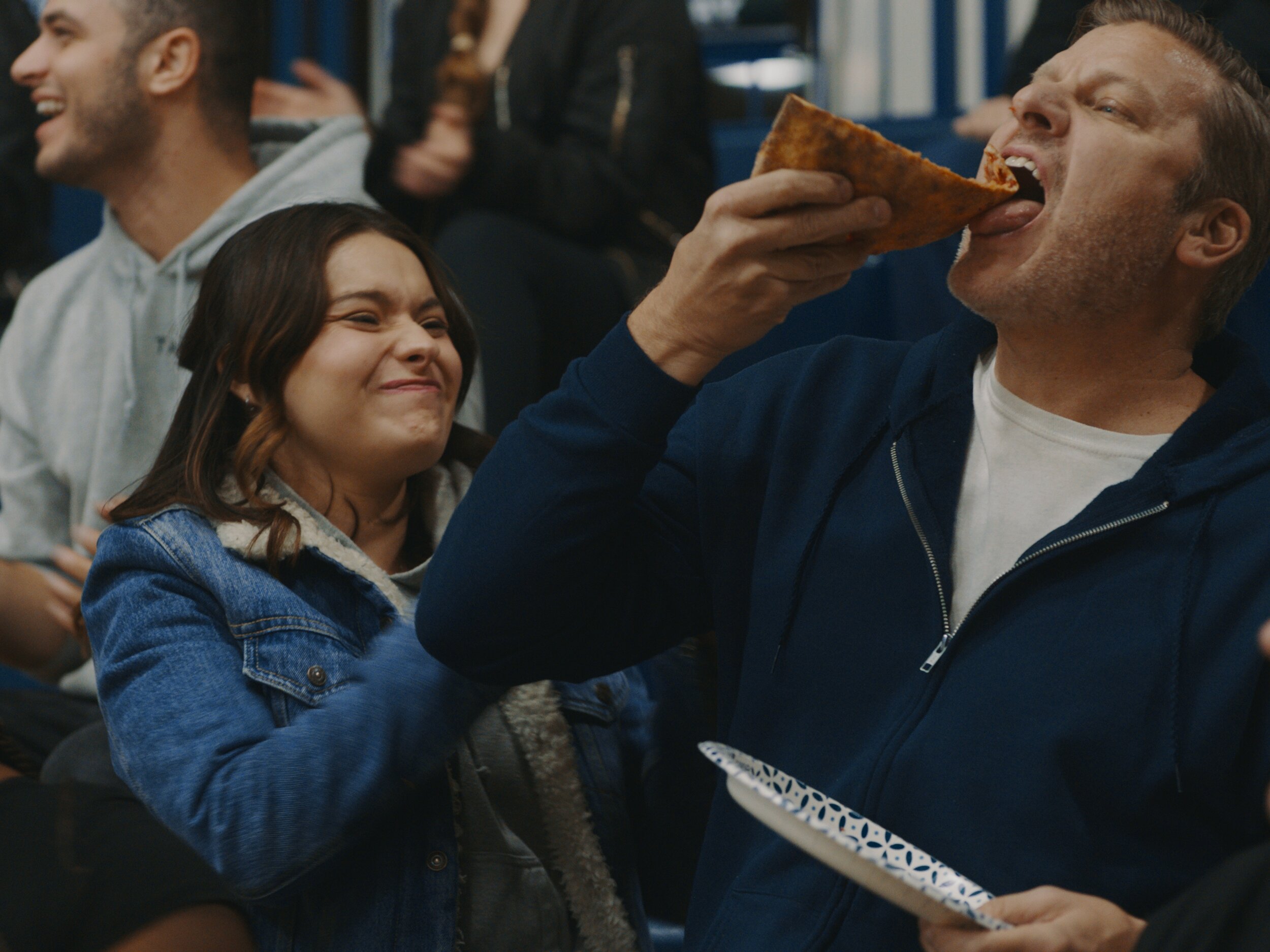 A man is eating a slice of pizza while a woman next to him grimaces.